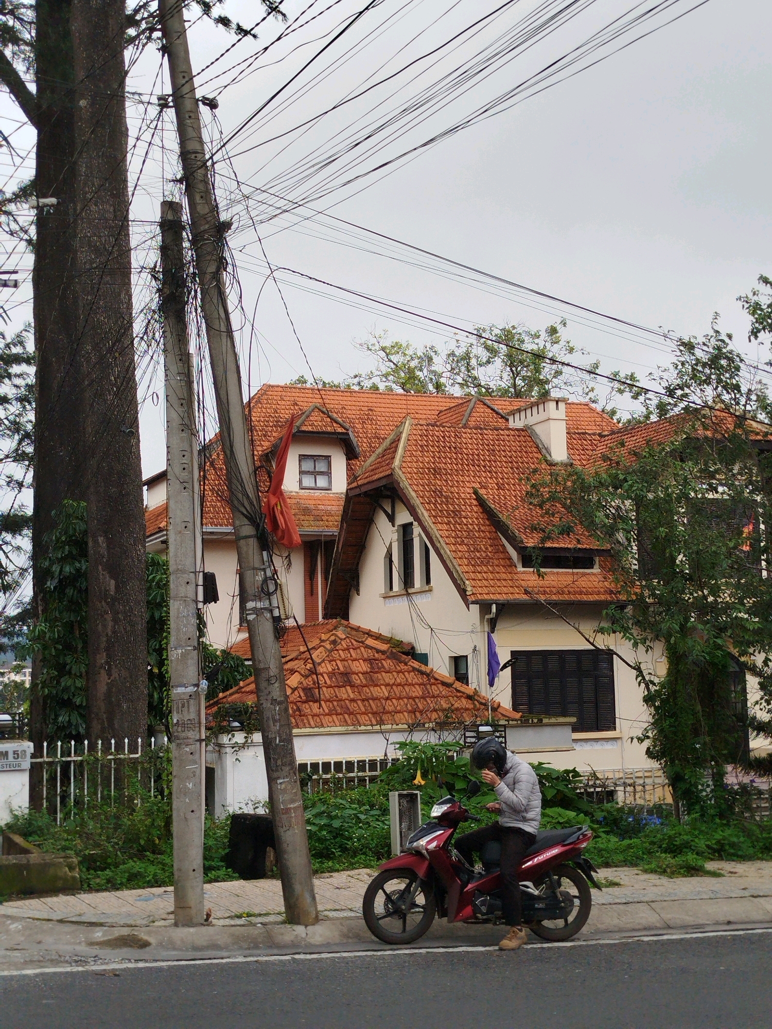 In Da Lat, Vietnam, a person wearing a light-colored puffer jacket and a dark helmet sits on a red motorcycle. The motorcycle is parked on the side of a paved road, near a sidewalk and a curb. Behind the motorcycle, a house with a distinctive orange-tiled roof and white walls stands. The house has several windows with dark shutters and a chimney. The scene is framed by a large tree to the left and a tangle of electrical wires overhead, supported by concrete poles. Lush green vegetation surrounds the house and lines the roadside. The sky is overcast.