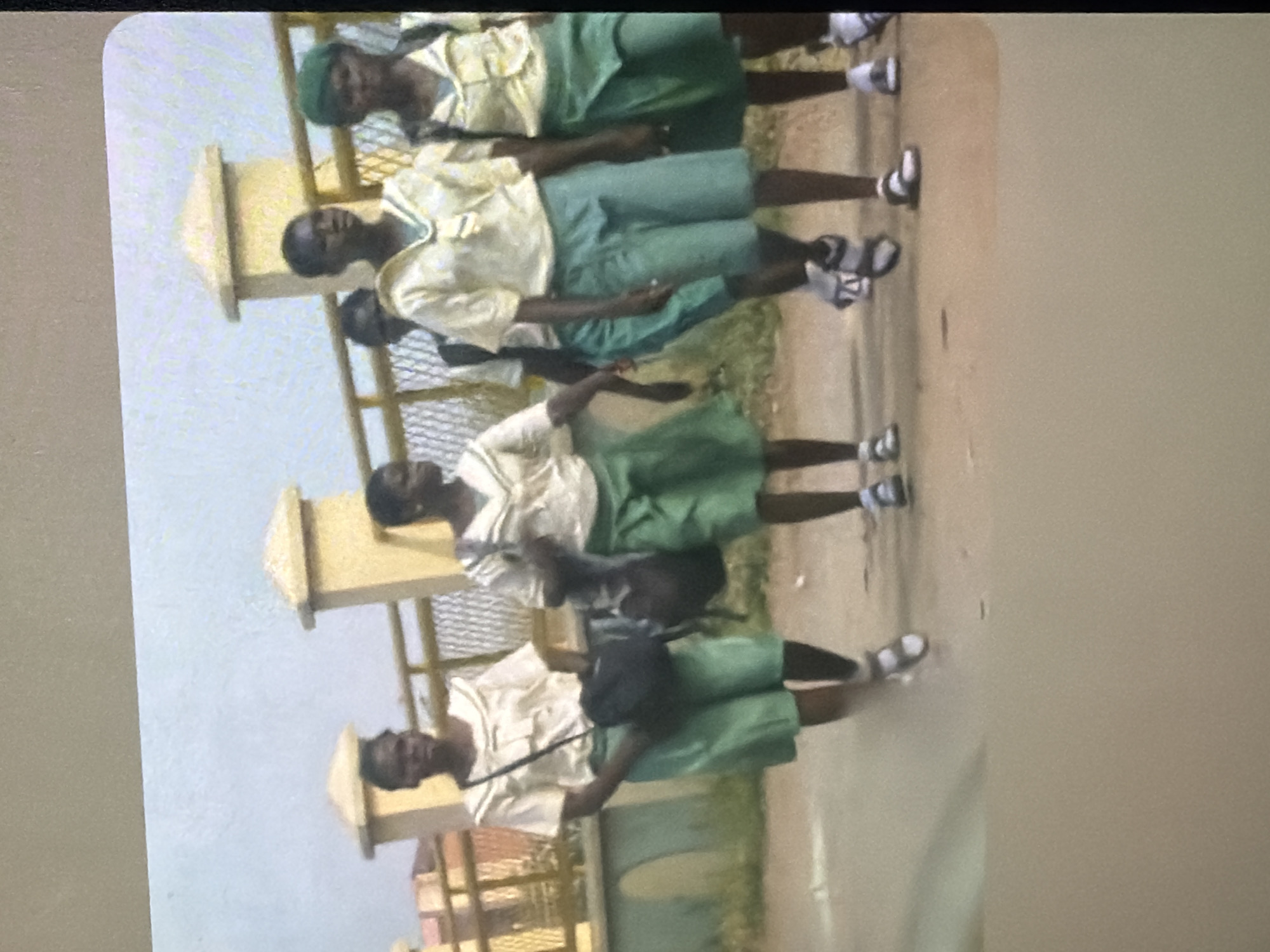 The image shows a group of five young Black girls in school uniforms walking on a paved surface in front of a fence and some buildings. They are all wearing light-colored blouses and green skirts. The girl on the far left is wearing a black bag over her shoulder and carrying another bag. The girl second from the right is wearing a green beret. The ground appears to be dirt or sand mixed with pavement. The sky is overcast and the lighting is diffused. The overall scene suggests an outdoor environment, possibly a school or a street near a school. The image is blurry and appears to be taken with a low-quality camera or at a distance.