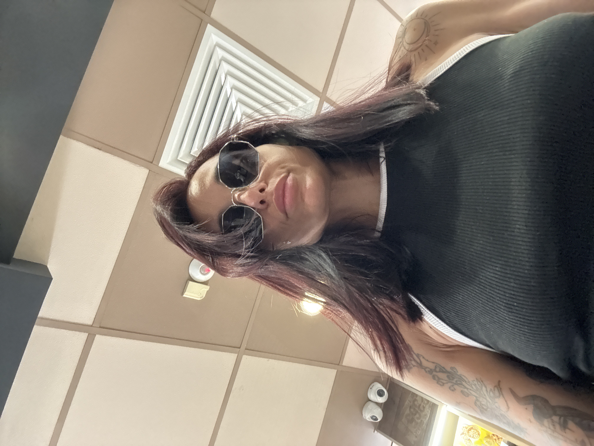 A low-angle, medium shot captures a woman wearing sunglasses and a black ribbed tank top with white trim. She has dark hair with reddish undertones. Her left arm, visible in the foreground, is adorned with tattoos. She is positioned indoors, with a tiled ceiling and a ventilation grate visible above her. To the left, two white security cameras are mounted on a wall. The background appears to be a retail or service environment, with shelves and products partially visible. The image does not provide any specific information or clues about the location being the city of Pleven, Bulgaria.