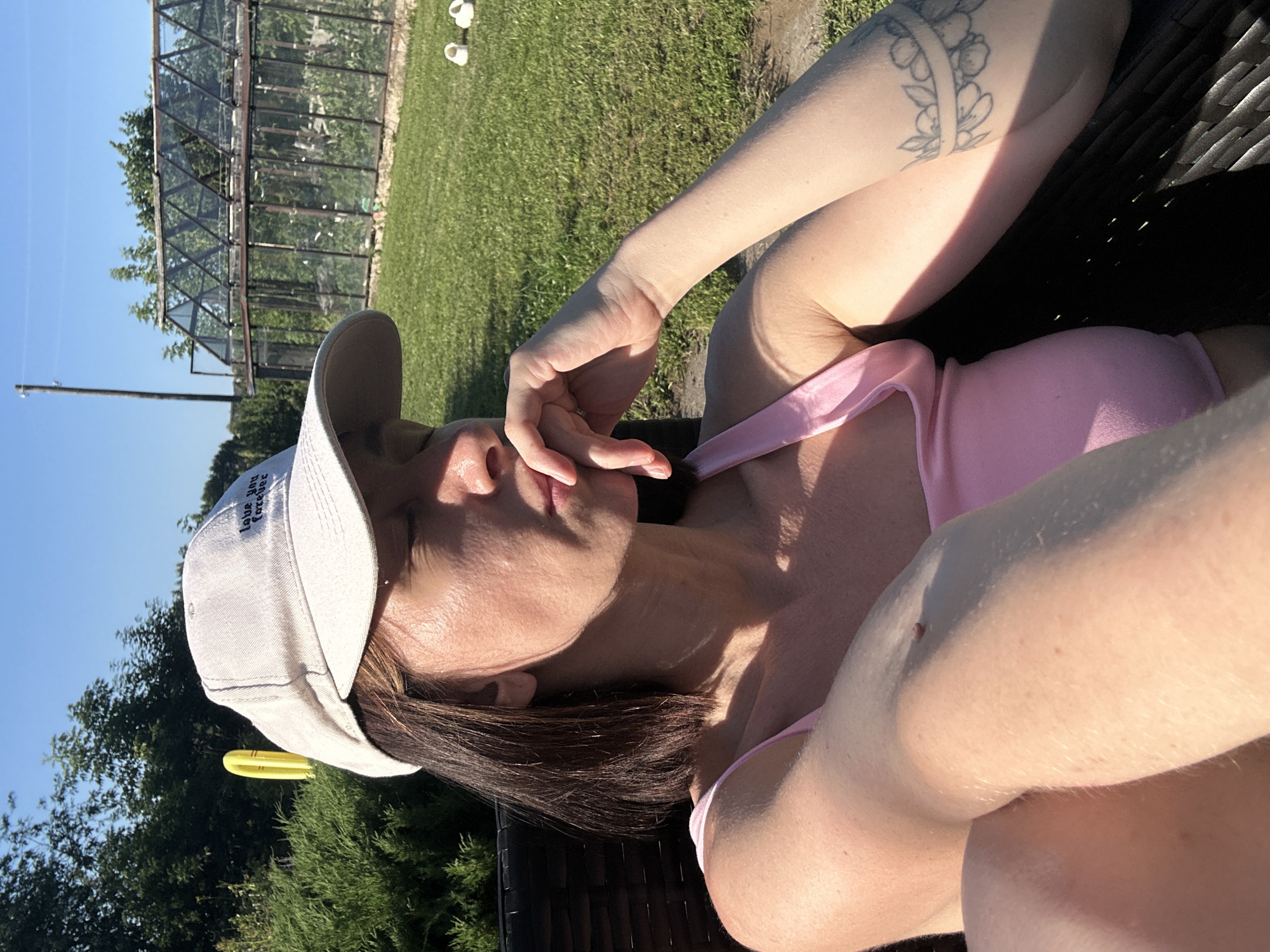 In the city of Galevo, Russia, a woman wearing a white baseball cap and a pink top is sitting outdoors. The cap has the text "love you forever" embroidered on it. She is leaning back in a dark-colored chair, with her eyes closed and her hand resting on her chin, as if lost in thought or enjoying the sunlight. Her left arm is visible, showing a tattoo on her forearm that appears to be a floral band. The background features a lush green lawn, a tall, dark green hedge, and a greenhouse with glass panes. The sky is a clear, bright blue, indicating a sunny day. The lighting suggests it is late afternoon, with shadows cast across the scene. The overall atmosphere is one of relaxation and peace.