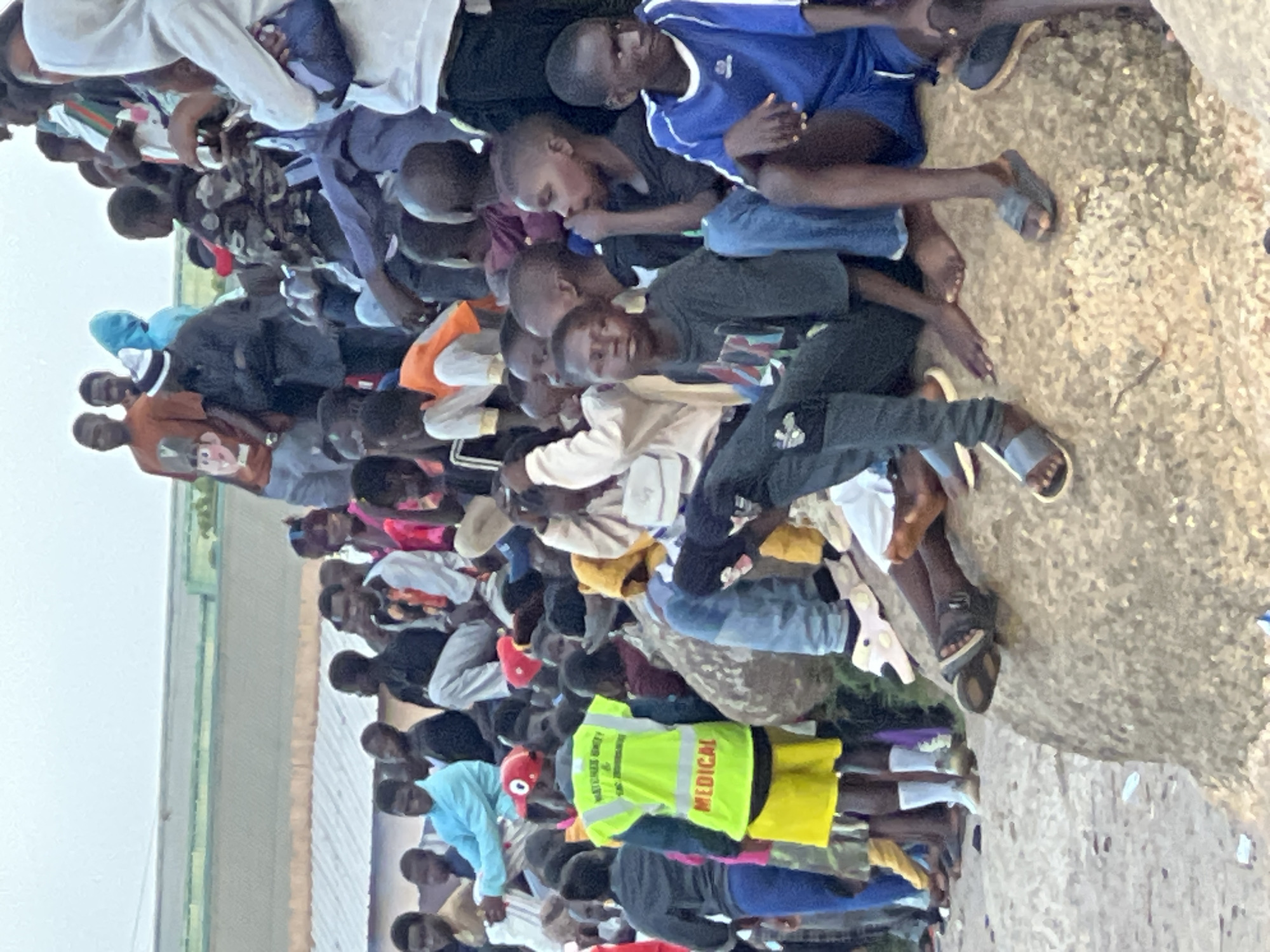 In the city of Jos, Nigeria, a crowd of people, predominantly young males, are gathered outdoors. The scene appears to be a public event or gathering, possibly at a stadium or an open-air venue, with tiered seating visible in the background. Many individuals are seated or standing, some looking towards an unseen focal point.
A prominent figure in the foreground is a young person wearing a bright yellow vest with "MEDICAL" printed in red, indicating a role in event safety or healthcare. This person is also wearing yellow shorts and a red cap. Surrounding this individual are other young people, some with arms around each other, suggesting a sense of community or shared experience.
The environment appears to be sandy or rocky, with loose debris on the ground. The lighting suggests it is daytime, possibly late afternoon or early evening, given the soft light. The background shows a large, light-colored structure with what looks like a corrugated roof. The overall atmosphere is one of a lively, communal gathering in a public space in Jos.
