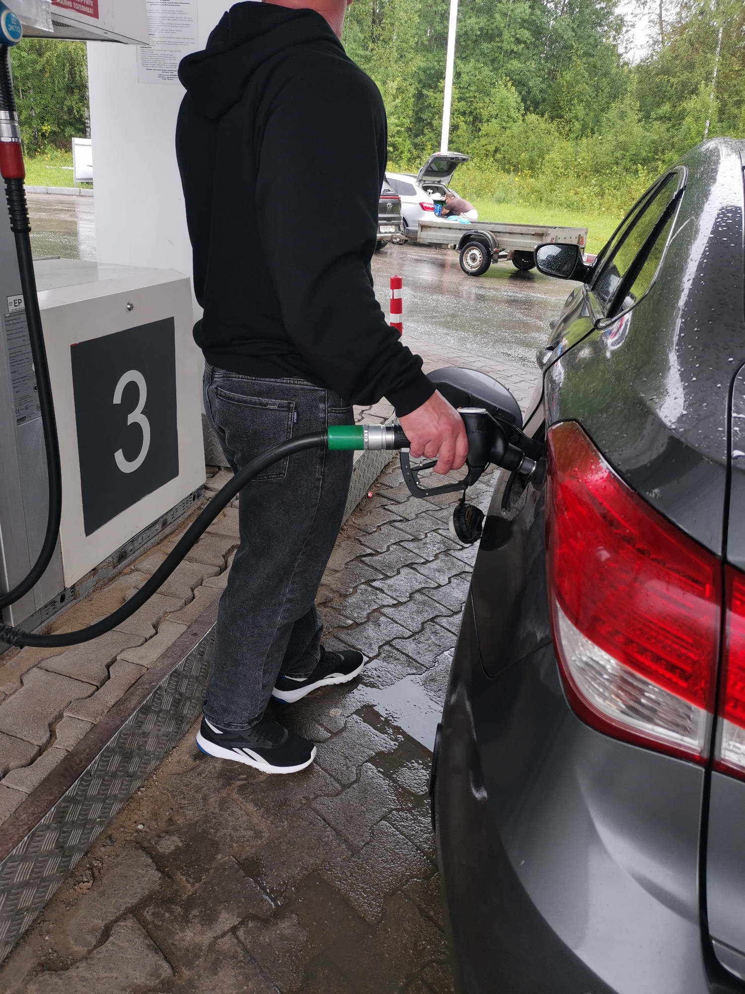 A person wearing a black hoodie and dark jeans is standing at a gas station, holding a fuel nozzle to the gas tank of a dark gray car. The nozzle is green and black and connected to a fuel pump labeled with the number "3". The ground is wet, with puddles of water on the brick pavement. In the background, another car is parked with its trunk open, and a trailer is attached to it. Trees and foliage form a backdrop to the scene. The weather appears overcast.