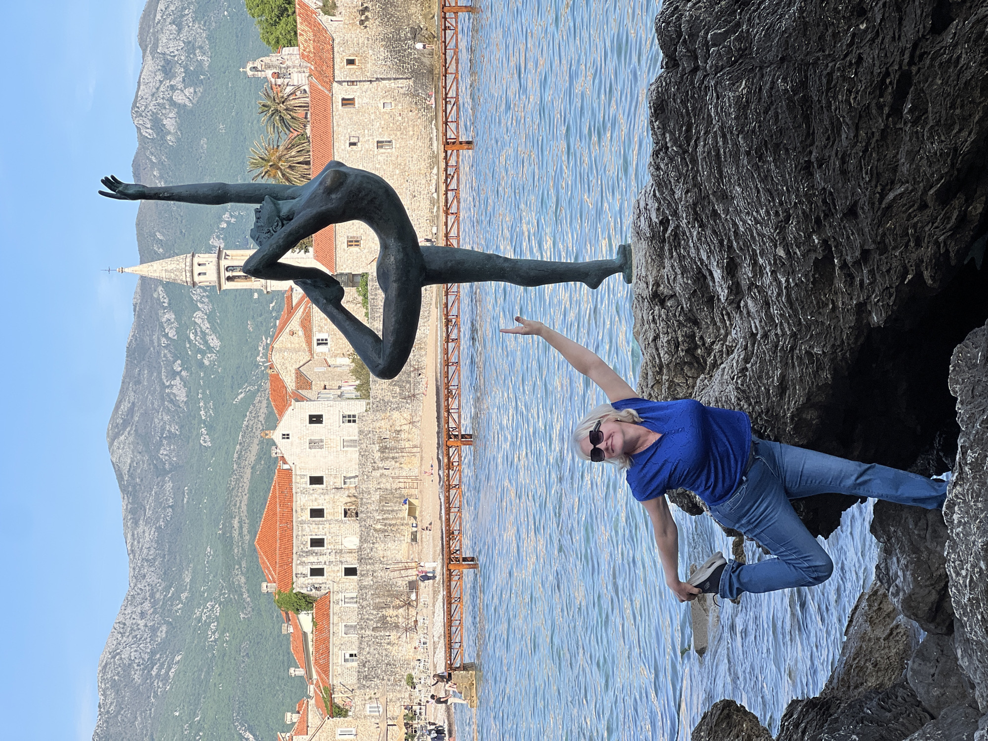 This outdoor, daylight photograph captures a scenic view along the coast of Budva, Montenegro, featuring a woman interacting with a famous statue.
In the foreground, on the right, a woman with blonde hair and sunglasses stands on a dark, rugged rock. She is wearing a blue short-sleeved top and blue jeans, and appears to be balancing or posing. Her left arm is extended upwards, mimicking the pose of a nearby statue, suggesting a playful or performative interaction.
To the left of the woman, situated on a separate rock closer to the water, is a striking bronze statue of a slender, nude female figure, commonly known as the "Dancer of Budva" or "Ballerina Statue." The statue is depicted in a dynamic and elegant pose, with one leg lifted and arms extended, conveying movement and grace.
The midground features the calm, blue waters of the Adriatic Sea, reflecting the sunlight. Behind the water and the statue, the historic Old Town of Budva is clearly visible. It consists of traditional stone buildings with distinctive orange-tiled roofs. A prominent church steeple rises above the town, and palm trees dot the landscape. A long, rusty-colored metal pier extends from the old town into the water.
In the background, majestic green and grey mountains rise under a clear, bright blue sky, indicating excellent weather conditions. The overall scene combines natural beauty, historical architecture, and art, with the woman engaging humorously with the iconic landmark. There is no visible text in the image.