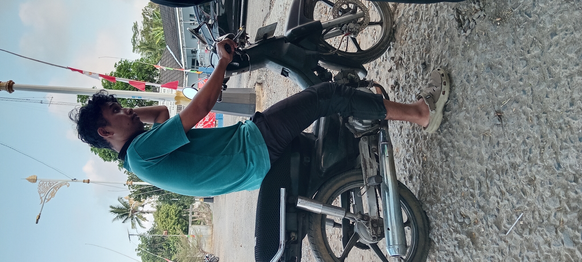 A young Indonesian man is sitting on a dark-colored motorcycle on a gravelly surface in Kalipucang, Indonesia. He is wearing a teal t-shirt and dark pants, with tan slip-on shoes. His left hand is raised to his ear, as if he is on the phone, and his gaze is downward. The motorcycle has a black seat and chrome accents, including a visible exhaust pipe. In the background, there are trees, buildings with tiled roofs, and a tall pole with red and white flags. The sky is clear blue with some white clouds.