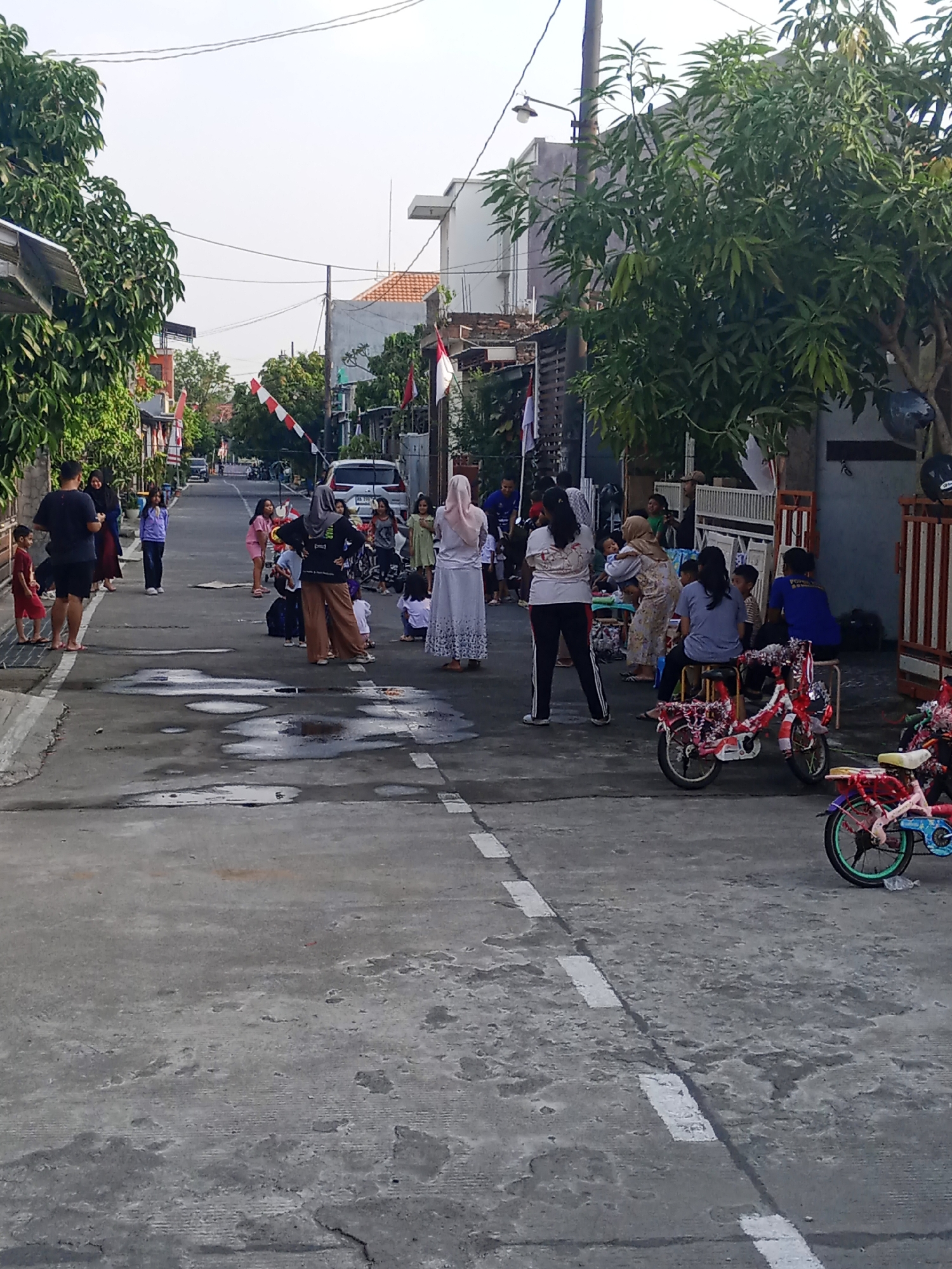 In the city of Demak, Indonesia, a street scene unfolds with a gathering of people, predominantly women and children, on a paved road. The atmosphere appears festive, with Indonesian flags, typically red and white, prominently displayed, suggesting a celebration or national holiday. Several individuals are standing and conversing, while others, particularly children, are seated or engaged in activities on the ground. Two bicycles, decorated with red and white streamers, are parked on the right side of the road, adding to the celebratory mood. A white car is visible further down the street, indicating that it is a residential area. The road surface is uneven with visible puddles of water, suggesting recent rain. Lush green trees line the sides of the street, providing a natural element to the urban environment.