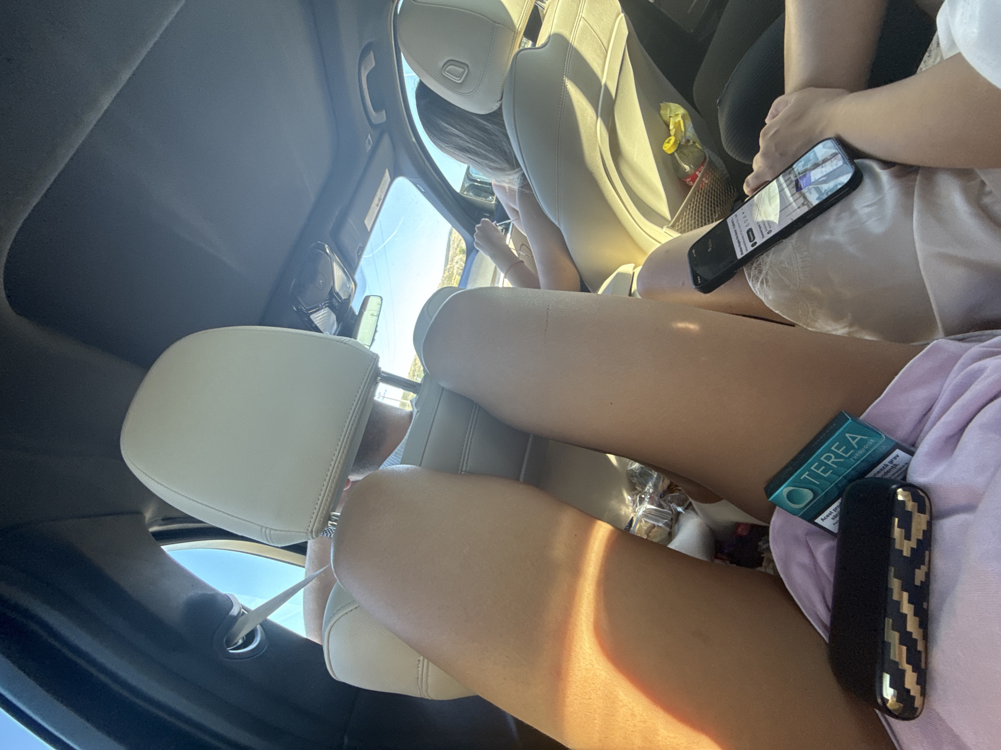 This image captures an interior view of a car, possibly in or near Popeni, Romania, on a sunny day. The foreground is dominated by the legs of at least two individuals. One person's bare legs are prominently displayed, positioned in the front passenger seat, with sunlight casting a warm glow. In the lap of this individual rests a black and gold patterned electronic cigarette holder and a teal-colored pack labeled "TEREA TURQUOISE". Also visible is a smartphone held by another person, displaying a YouTube video. The car's interior features light-colored leather seats and a sunroof. In the background, a person appears to be driving. The exterior scenery, visible through the car windows, suggests a rural or suburban landscape with low hills and clear blue skies. A mesh organizer pocket in the back seat holds a bottle of water and a snack bag.