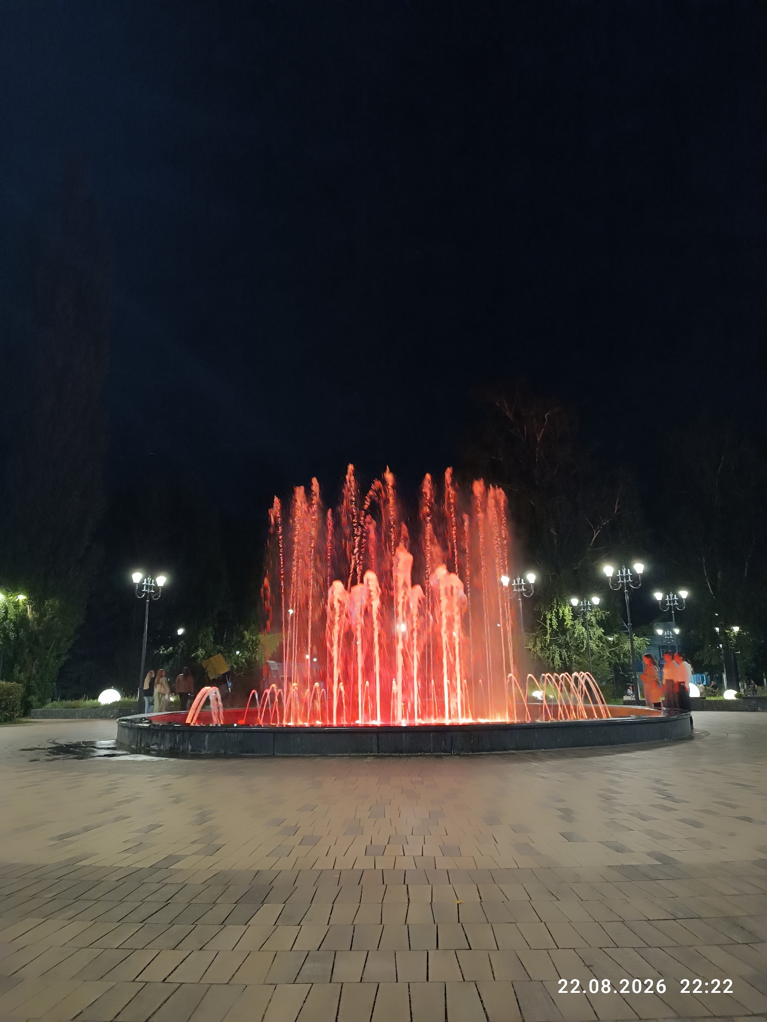 A vibrant, illuminated fountain is the central focus of this nighttime scene. The water jets are lit with a strong red glow, creating a dramatic visual effect. The fountain is situated in a paved public area, likely a park or square, with trees and lampposts visible in the background. Several individuals are present, appearing to be casually walking or standing near the fountain. The ground is paved with interlocking rectangular stones. In the lower right corner, a timestamp indicates "22.08.2026 22:22". While the image captures a specific moment, it doesn't explicitly identify the city of Sterlitamak, Russia.