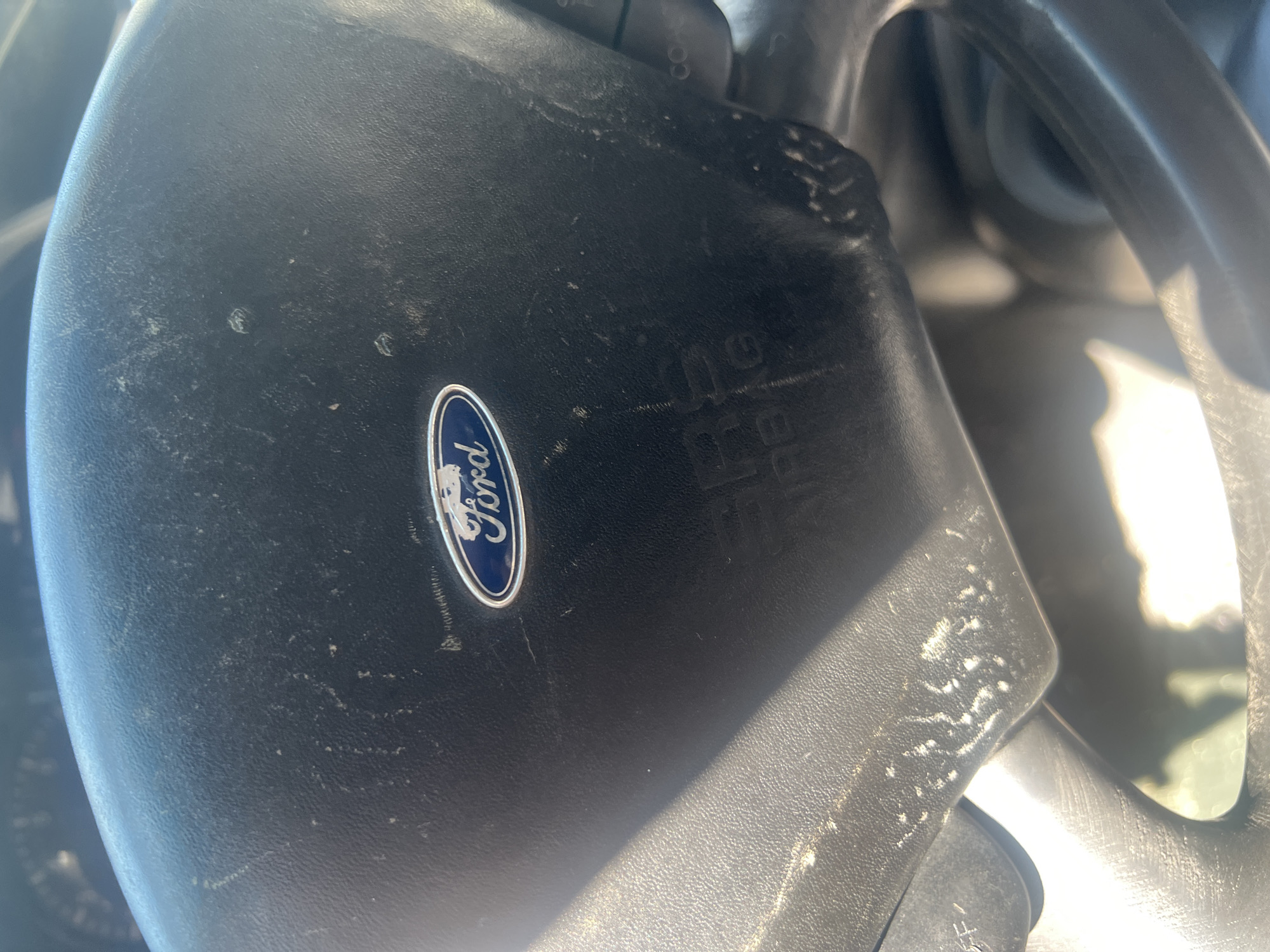 The image shows a close-up of a steering wheel with the Ford logo prominently displayed in the center. The steering wheel appears to be made of black plastic and is somewhat dusty and worn, with visible scratches and scuff marks. Below the Ford logo, the letters "SRS AIRBAG" are embossed into the plastic. The lighting suggests it is daytime, with sunlight casting a glare on the left side of the steering wheel. The background is blurred, but it appears to be the interior of a vehicle. There is no information available to determine the location in the city of Unknown, Unknown.