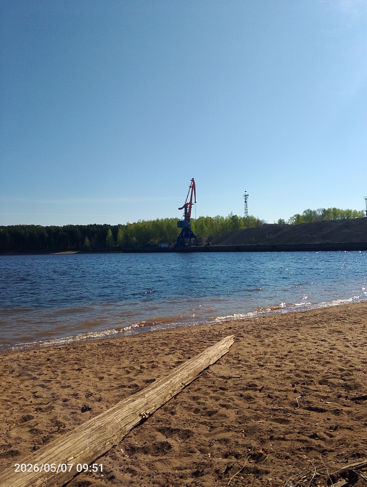 The image depicts a sandy beach on the bank of the Kama River in Chaikovsky, Russia. In the background, across the water, is a port facility with a crane and other industrial structures. The sky is clear and blue. A piece of driftwood lies on the sand in the foreground. The lower left corner of the image contains a date and time stamp indicating "2026/05/07 09:51."