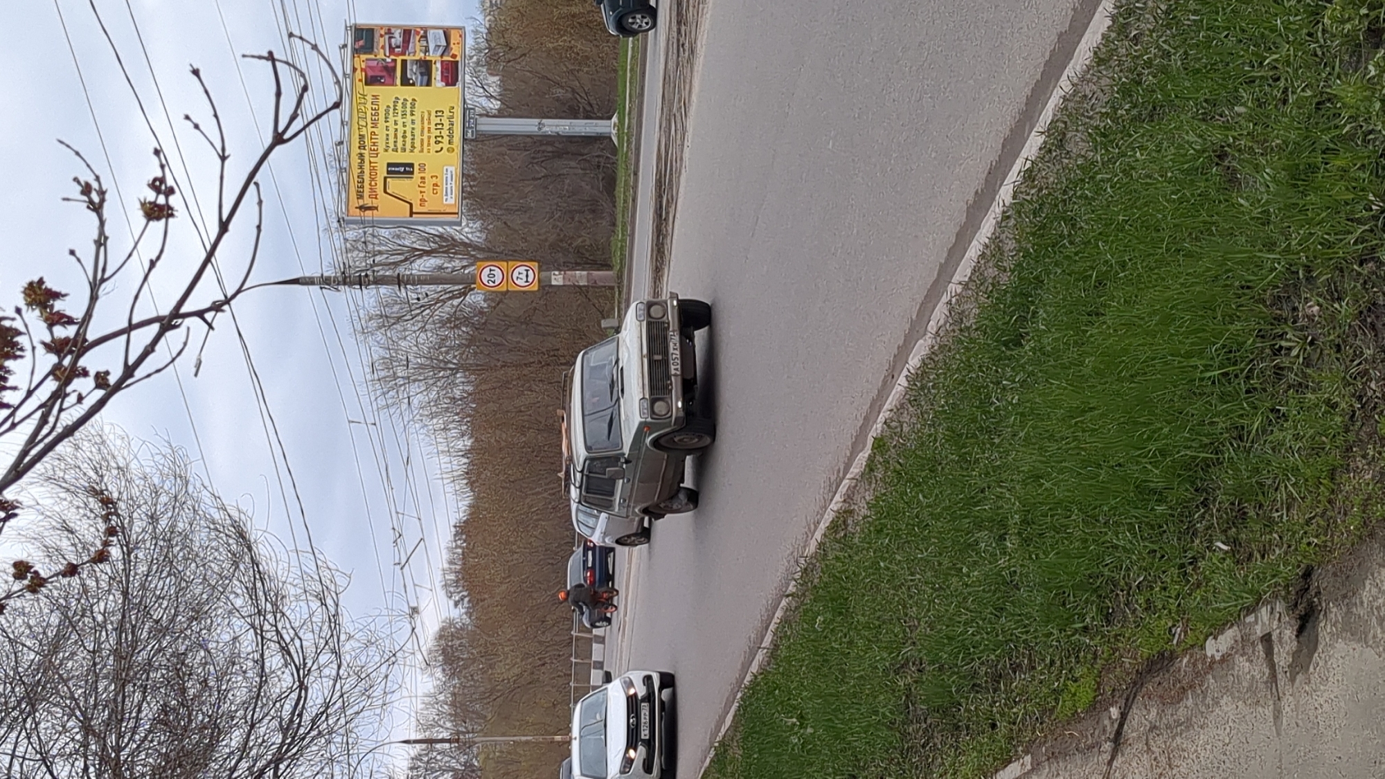 The image shows a street scene in Ulyanovsk, Russia. A silver Lada Niva is driving on the road, with other cars behind it. There is a motorcyclist present. The surroundings are primarily residential, with trees lining the street. A large billboard advertises a furniture store called "Mebelny Dom Charlie" (Мебельный Дом "CAPAC"), with a phone number 93-13-13 and website mdcharli.ru. The billboard also features advertised prices for kitchens, sofas, wardrobes, and beds. There are also street signs indicating weight restrictions.