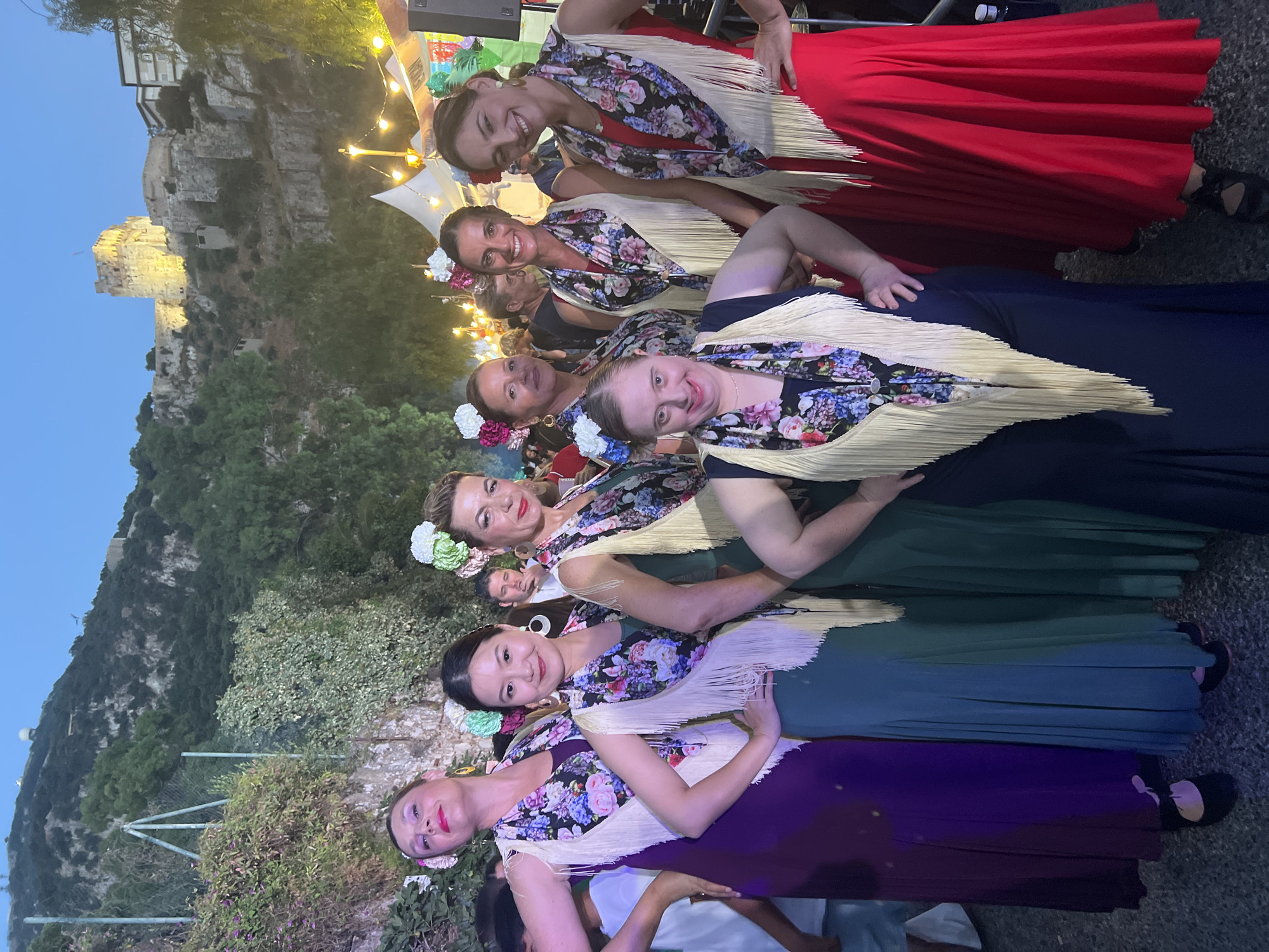 The image depicts a group of seven women posing outdoors in Gibraltar. In the background, the Rock of Gibraltar is visible, with the Moorish Castle illuminated on its summit. The women are dressed in brightly colored, floor-length dresses and are adorned with floral headpieces and fringed shawls, suggesting a cultural or celebratory event. One woman in the front row has Down syndrome. They appear to be participating in a festival or performance. The lighting suggests it is either dusk or early evening. Tents and decorative lights are visible behind the group, further indicating a festive atmosphere.
