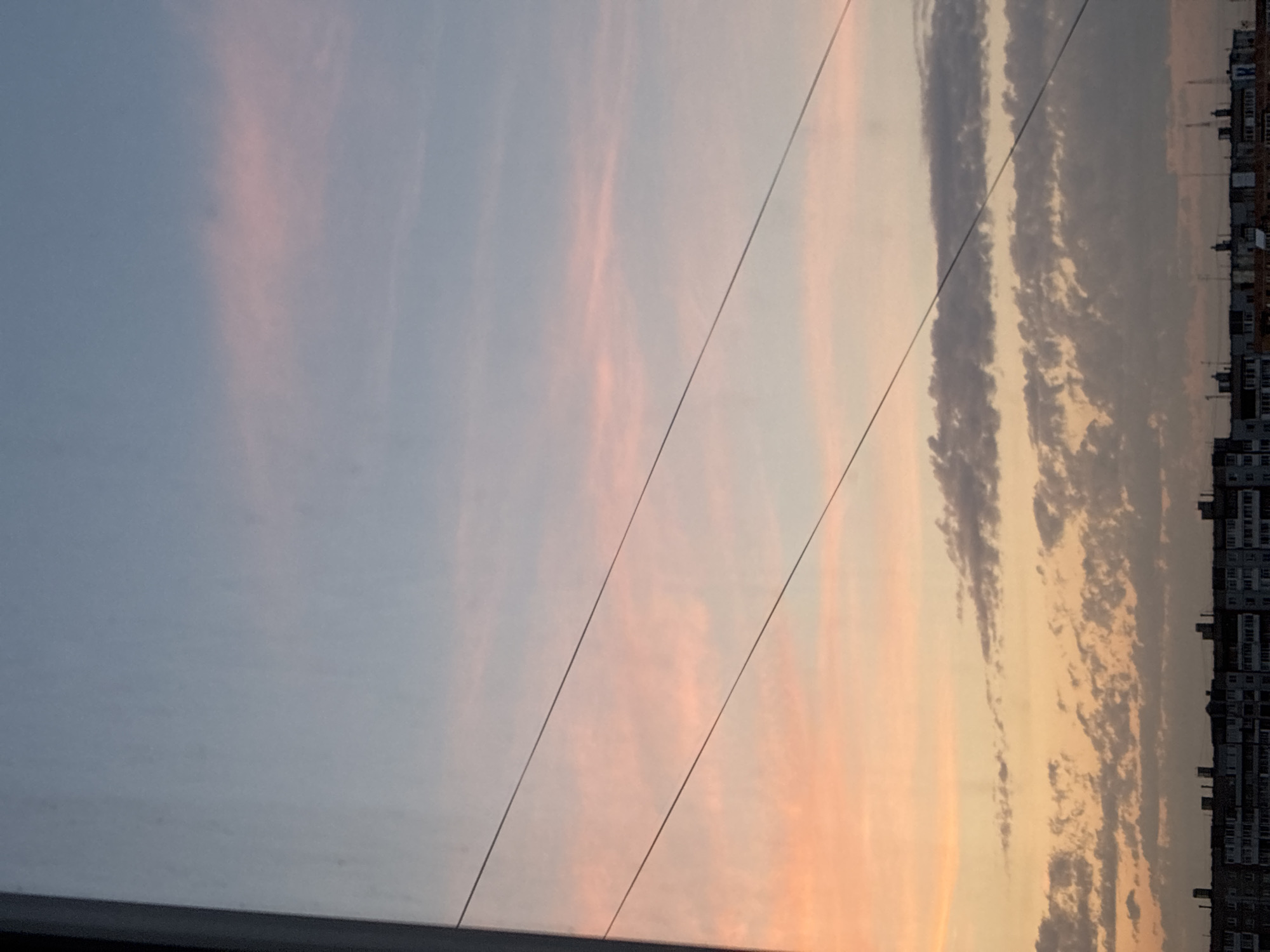 The image shows a cloudy sky at sunset over a city. The sky is a gradient of colors, from pale blue at the top to orange and pink near the horizon. There are several thin, dark wires stretched across the sky. Below the sky, there is a silhouetted row of apartment buildings. The buildings are dark and rectangular, with many windows visible. The overall scene is one of a typical urban sunset.