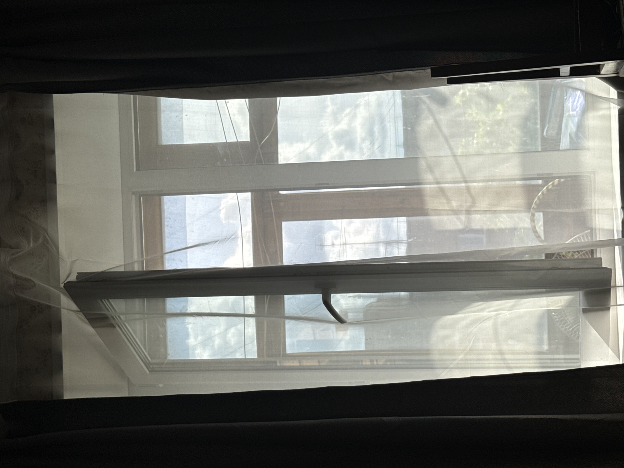 The image shows a partially opened window with a sheer curtain obscuring the view. The window is divided into multiple panes. Outside, the sky is visible, and some indistinct foliage can be seen. The window's handle is visible in the open section. The image is taken from a darkened room, with dark fabric or curtains framing the window. It is impossible to ascertain the location of this image to Mytishchi, Russia.