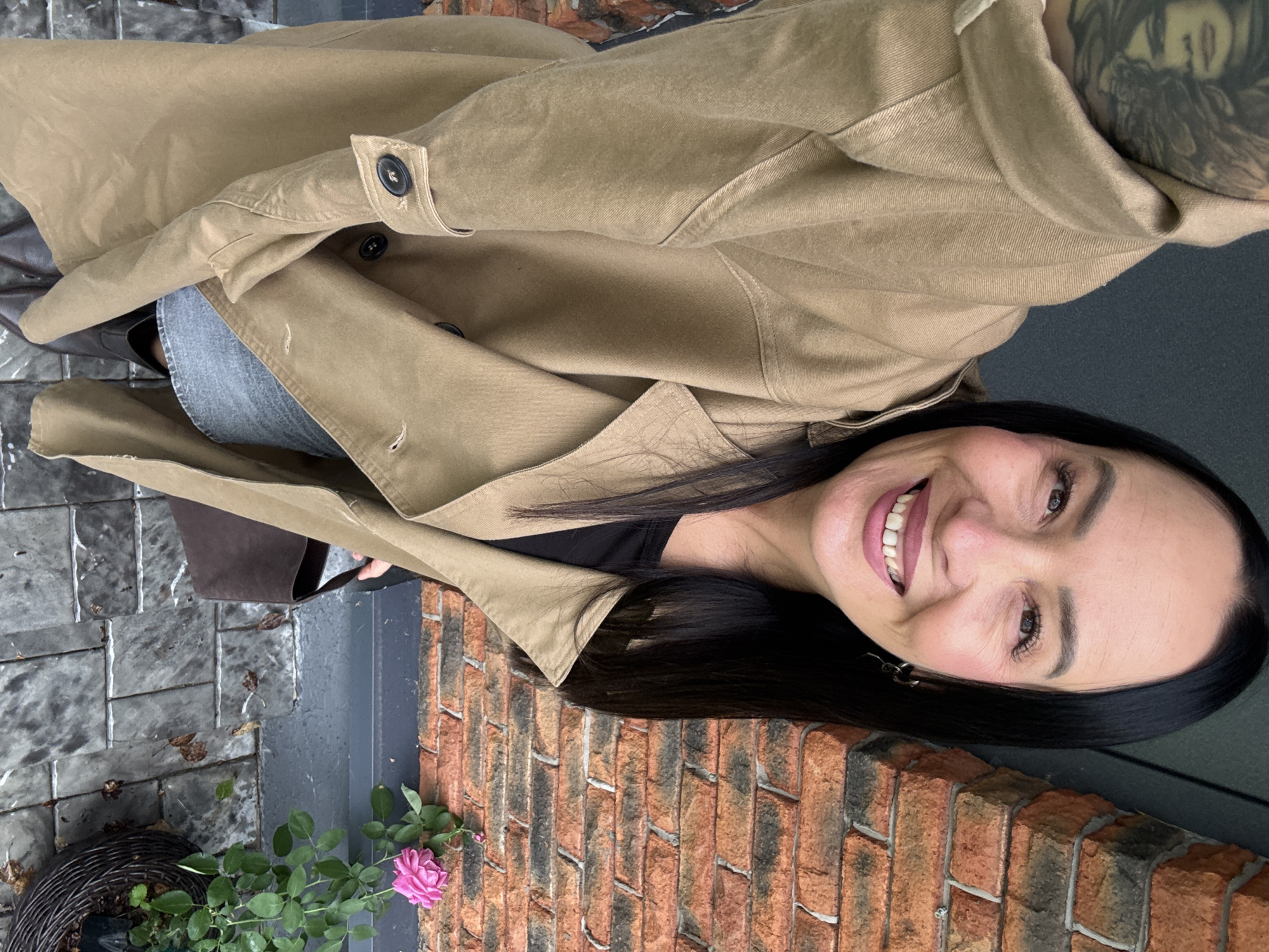 A woman is captured in a selfie, standing outdoors next to a brick wall. She is wearing a tan trench coat, a black top, and a denim skirt. A brown handbag is visible. To her right, a pink rose is in bloom in a woven basket. The ground is paved with gray tiles. The background shows a brick wall and a dark wall. The image was taken on a cloudy day.