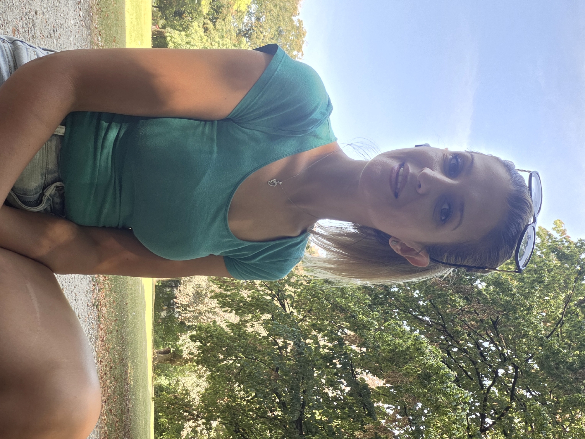 A woman with blonde hair pulled back in a ponytail and sunglasses perched on her head is captured in a medium shot, seated outdoors in a park. She is wearing a teal t-shirt and denim shorts, and a silver necklace with a heart-shaped pendant. The background features a large, leafy tree on the right side of the frame, with a bright blue sky above. The ground in front of her is a gravel path. The overall impression is of a relaxed, sunny day in a natural setting. While the specific location in Munich, Germany is not explicitly stated, the scene suggests a park within the city.