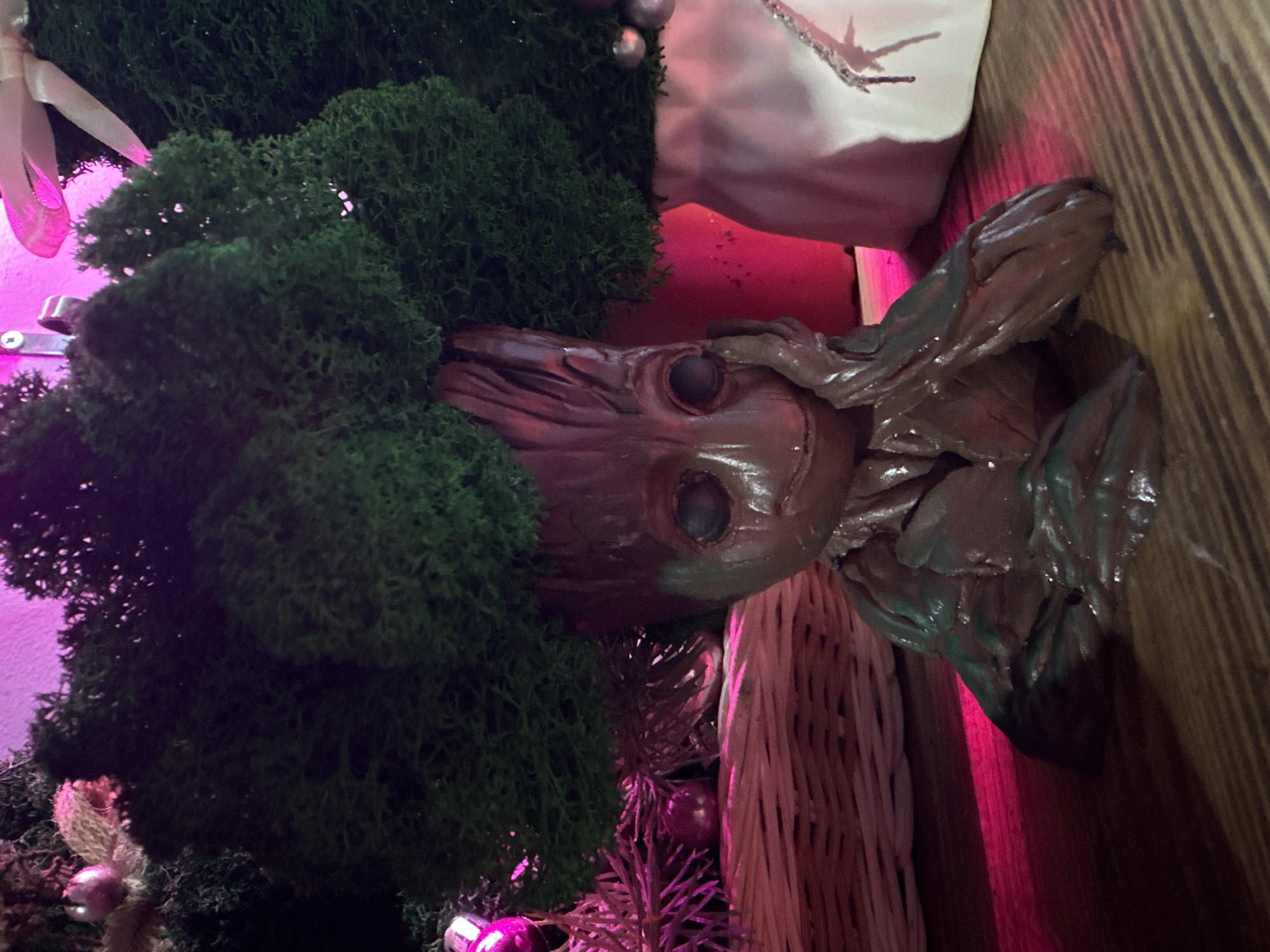 An indoor scene displays a brown, wood-textured figurine resembling the character "Groot," featuring a smiling expression and dark eye sockets. The figurine's left hand rests against its left cheek. A dense mass of green preserved moss forms the top of its head. The figure is situated on a light brown wooden surface exhibiting horizontal grain patterns. To the left background, a light-colored woven basket contains several pink spherical ornaments and thin decorative branches. To the right background, a red cylindrical pot is partially visible behind the figurine, adjacent to a white ceramic vase with silver-colored decorative streaks, which also contains green moss. The environment is illuminated by a prominent pink-purple light source from the upper left, casting a magenta glow on the background wall and objects. The image does not contain identifiable features to determine a specific location within Rostov-on-Don, Russia.