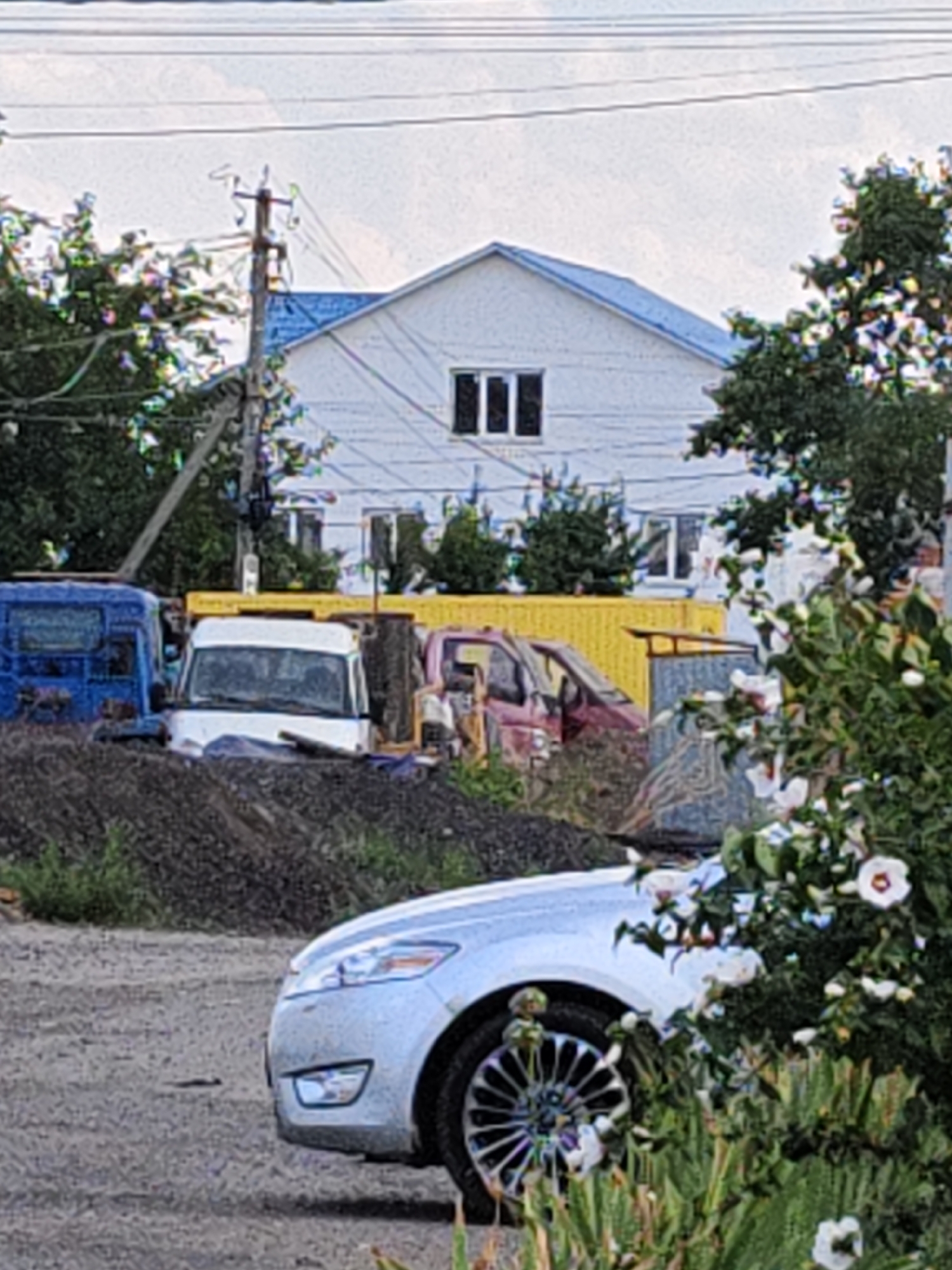 The image captures a cluttered outdoor scene with a white house featuring a blue roof in the background. In the foreground, a silver car is parked on a dirt road, with part of its wheel and front bumper visible. To the left of the car, a pile of dark soil or gravel is present. Behind this, several vehicles are visible, including a blue van, a white van, and a red van, positioned near a bright yellow structure. The environment appears to be a residential or industrial area with trees and power lines present. No individuals are depicted in the image.