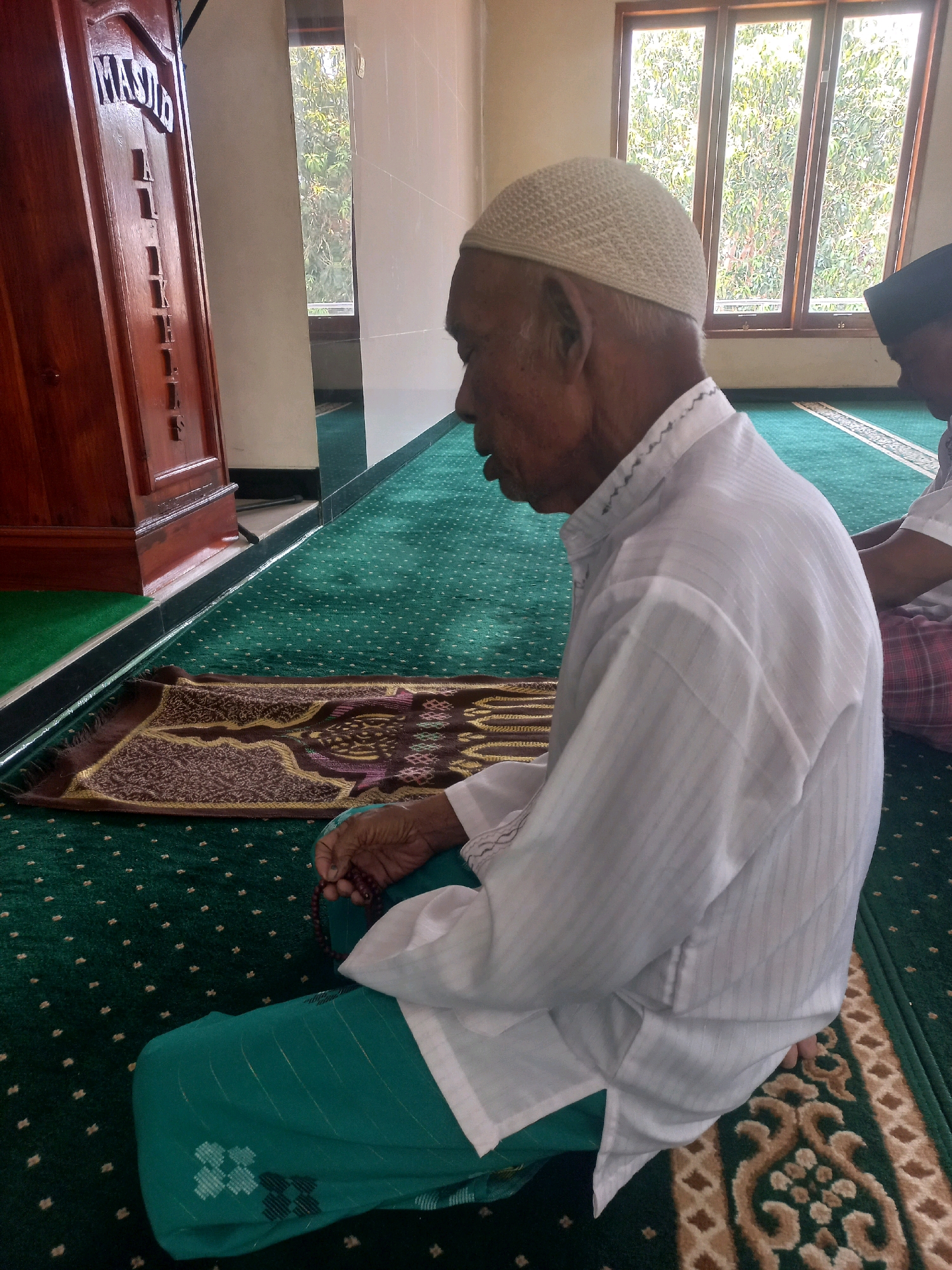 In a room with a green carpet and large windows, an elderly man in a white shirt and a white skullcap is sitting on the floor. He is facing to the right and appears to be praying. He is holding prayer beads in his right hand. To his left, a wooden cabinet with Arabic writing on it is visible. To his right, another person is partially visible, wearing a black skullcap and a white shirt. The room appears to be a mosque. The windows show trees and foliage outside, suggesting a location in Nguntoronadi, Indonesia.
