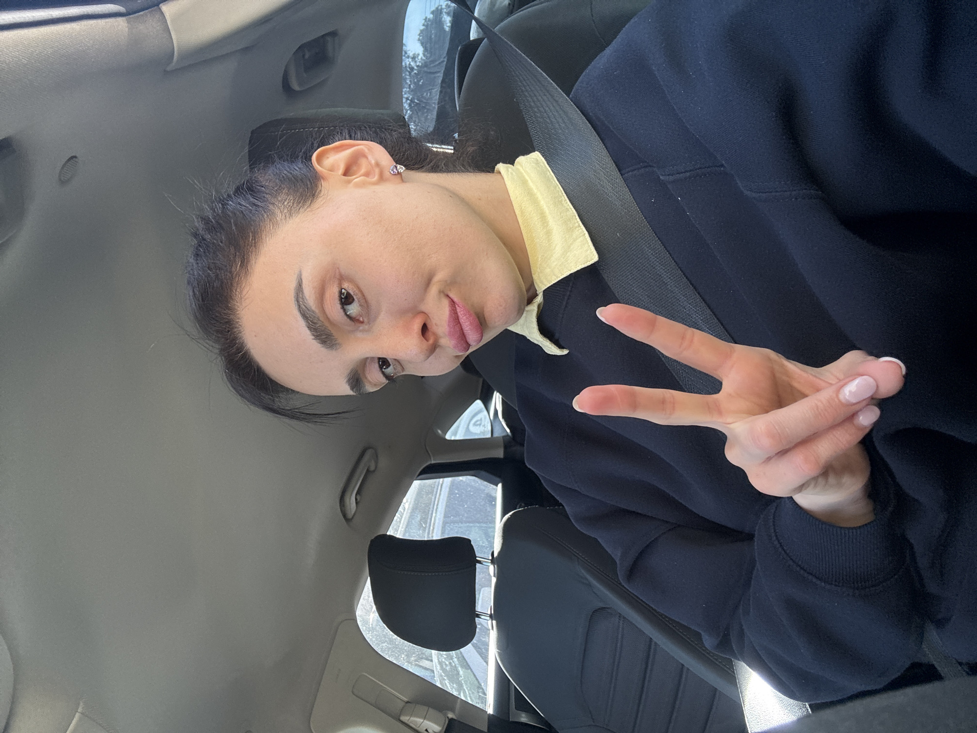 A woman sits in the driver's seat of a car, wearing a seatbelt. She has dark hair pulled back, light skin, and is making a peace sign with her right hand. She is wearing a dark blue sweatshirt over a collared yellow shirt. The car's interior is a light gray color. The car's headrest is visible, along with a glimpse of the outside through the car window. There is no specific detail to determine a location in Tbilisi, Georgia.