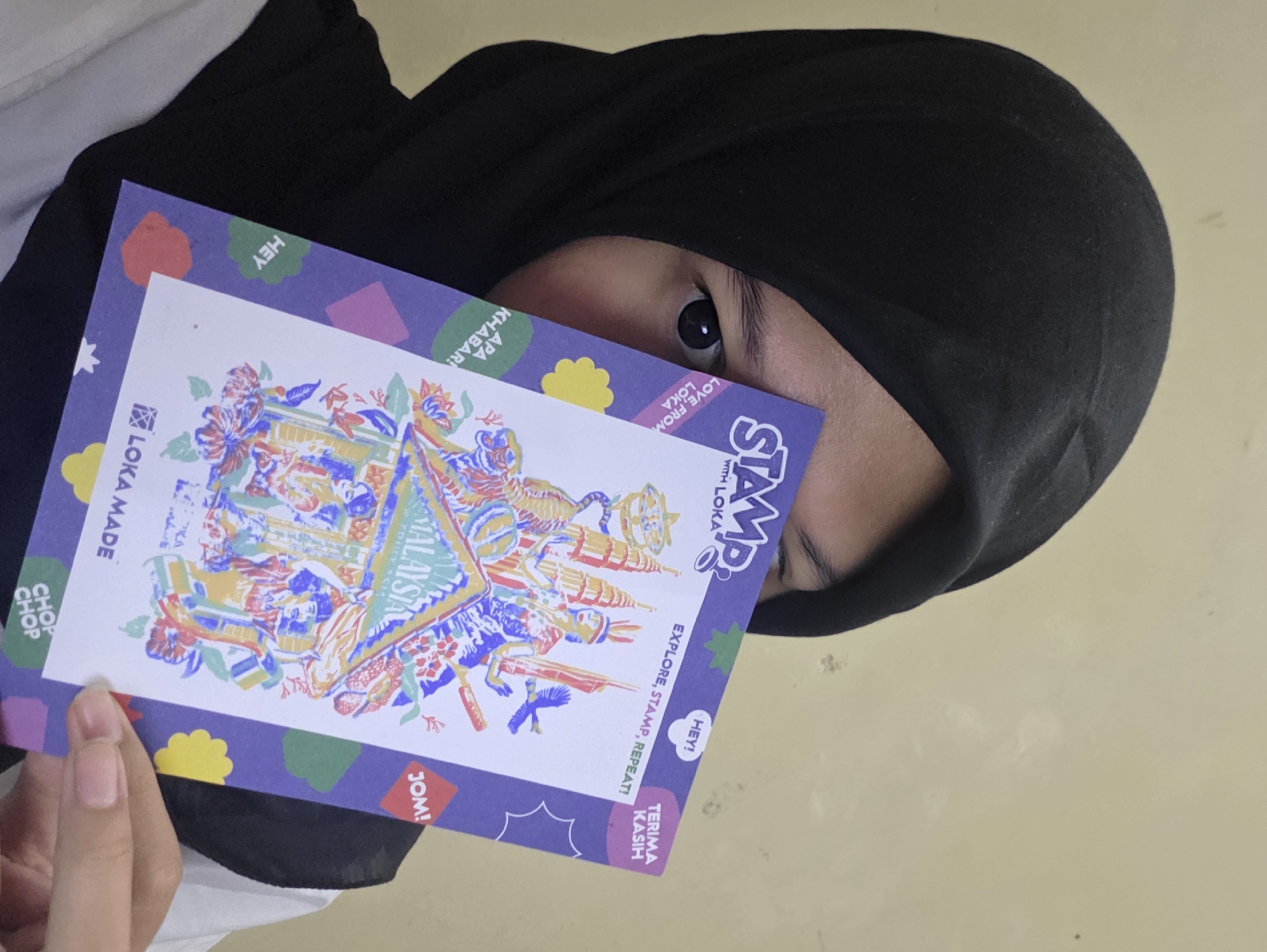 A person wearing a black hijab is holding a card with colorful designs and text. The card reads "STAMP with LOKA", "EXPLORE, STAMP, REPEAT!", "LOKA MADE", and mentions "MALAYSIA". The card is partially covering the person's face, with one eye visible. The background is a plain wall.