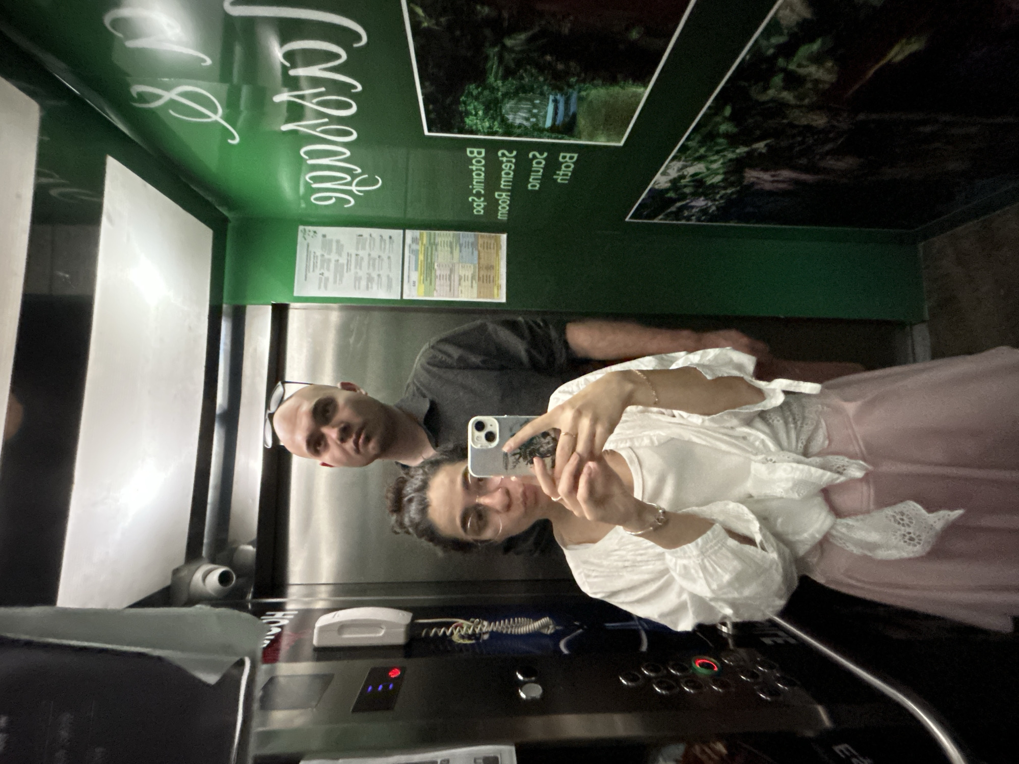 A selfie captured inside an elevator shows a woman in the foreground taking the photo with a smartphone, alongside a man standing slightly behind her. The image is rotated 90 degrees counter-clockwise. The woman, with dark hair and glasses, is wearing a white ruffled top and a light pink skirt. She has a watch or bracelet on her left wrist and rings on her fingers. The man, bald and with glasses pushed up onto his head, wears a dark grey short-sleeved shirt. Both individuals look towards the camera with neutral to slightly smiling expressions, appearing relaxed.
The elevator interior features reflective metallic panels on one side, where the individuals are visible, and dark green panels on the other side. The green panels are adorned with white, flowing script text and images depicting lush green foliage, suggesting a natural or spa-like theme. Visible text on the green wall, partially reflected, includes "Spa & Massage" (with the first word partially obscured, possibly "Caspas"), and mirrored text like "Sauna," "Steam Room," and "Botanic Spa." Two smaller, rectangular information sheets, likely containing schedules or service details, are also affixed to the green wall, though their text is illegible.
The elevator control panel is visible below the individuals, featuring an array of buttons and an illuminated red light, possibly indicating an active function or call. A white intercom handset is mounted to the left of the control panel, and a small security camera or sensor is positioned above it. The scene captures a casual moment of travel, likely within a hotel, spa, or wellness center in Esenler, Turkey, given the provided location context. The lighting is internal, so no time of day or weather details are discernible.