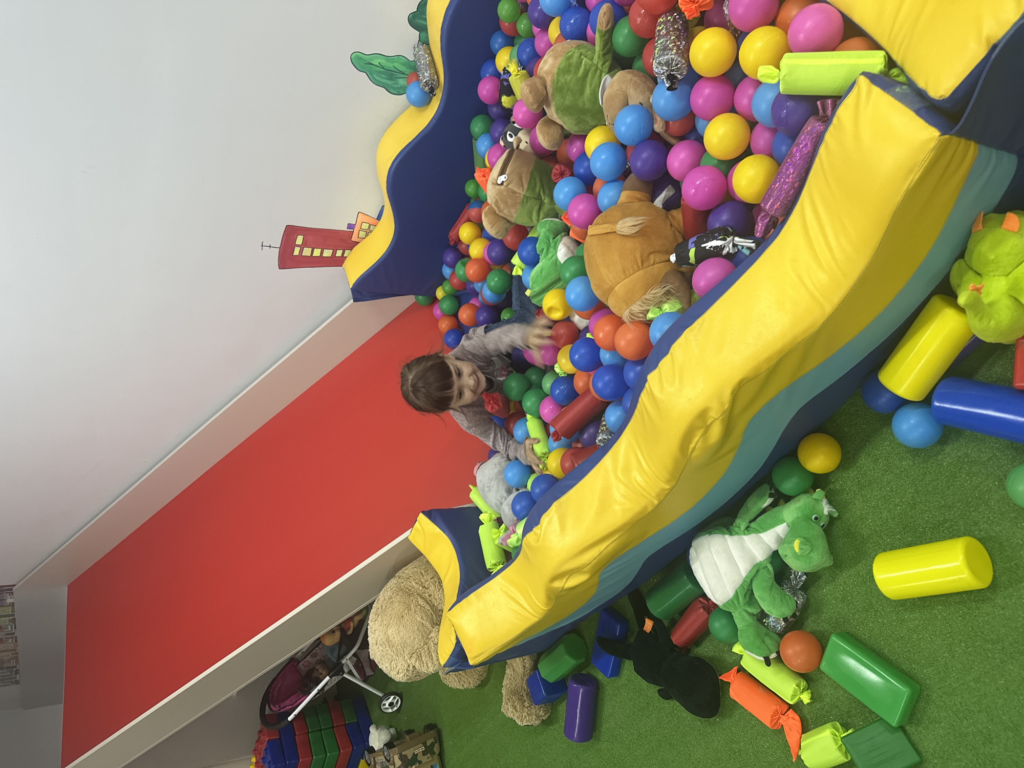 A young child, appearing to be a girl with brown hair, is shown joyfully playing in a large, colorful ball pit. Her arms are outstretched as she interacts with the multitude of plastic balls, and she has a happy, engaged expression.
The ball pit is a prominent feature of an indoor play area, constructed with soft, padded sections in bright yellow and blue. It is densely packed with plastic balls of various colors, including blue, pink, yellow, orange, and purple. Interspersed among the balls are several large, plush stuffed animals, such as brown monkeys or bears, and a green dinosaur or dragon, adding to the playful environment. Some shiny, tube-like objects are also visible within the balls.
The setting appears to be a dedicated children's play space, possibly a soft play center, daycare, or a well-equipped home playroom. One wall is painted a vivid red, while an adjacent white wall features a charming mural of cartoon-style buildings in orange and red with yellow windows, suggesting a city theme. The floor surrounding the ball pit is covered with a green carpet or mat, resembling artificial grass. On this green surface, various soft play shapes in green, yellow, blue, and purple are scattered, along with more stuffed animals, what looks like a toy stroller, and colorful building blocks.
The image is brightly lit, indicative of an indoor setting. The vibrant colors of the balls, soft play structures, and wall decor contribute to a cheerful and stimulating atmosphere. There is no visible text in the image.