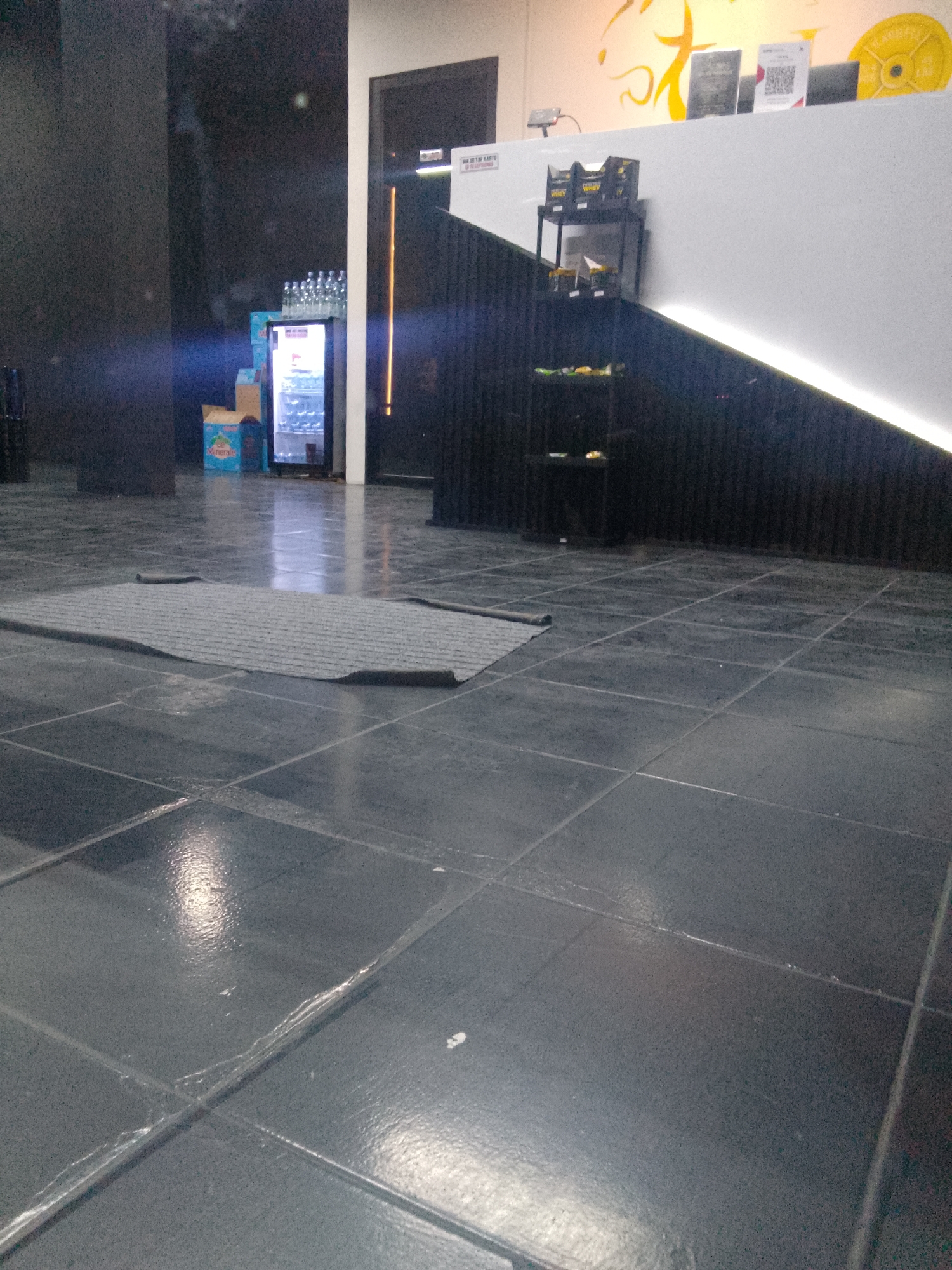 The image shows the interior of a gym or fitness center in Trenggalek, Indonesia. In the foreground, a gray mat is laid out on a dark, tiled floor. To the left and slightly behind the mat is a small, illuminated cooler containing bottles. Adjacent to the cooler are cardboard boxes, one of which has a blue label with white text that appears to read "Minerals." Further back, a counter or reception area is visible, with shelves displaying boxes of what appear to be protein supplements, along with various signage and equipment. The overall lighting suggests an indoor environment, possibly with artificial light sources contributing to the illumination. The gym appears to be equipped for various fitness activities.