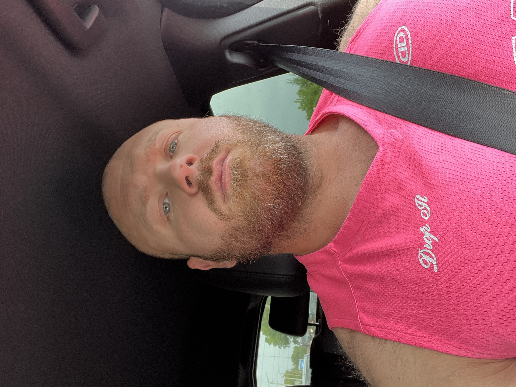A man is seated in a car, wearing a pink athletic shirt with the phrase "Drop It" printed on it. He is wearing a black seatbelt. His face is visible, with a short beard. The exterior view through the car window shows trees, buildings, and a street. The image was not taken in Chaikovsky, Russia, so that detail cannot be verified.