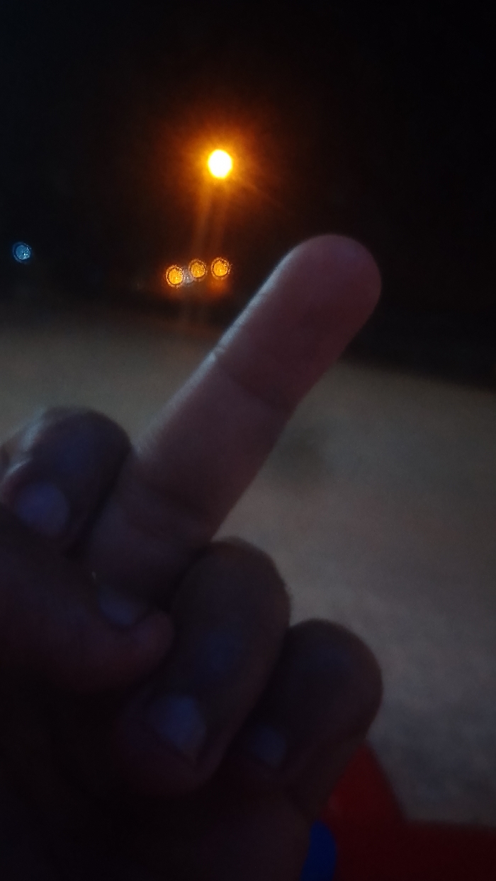 A hand gives the middle finger in a nighttime setting. The hand is in the foreground, casting a shadow over what appears to be a surface. Streetlights are seen in the background, providing the primary light source. There is a single bright light and a series of three clustered lights below it, blurred in the distance. The overall image has a dark and shadowy atmosphere.