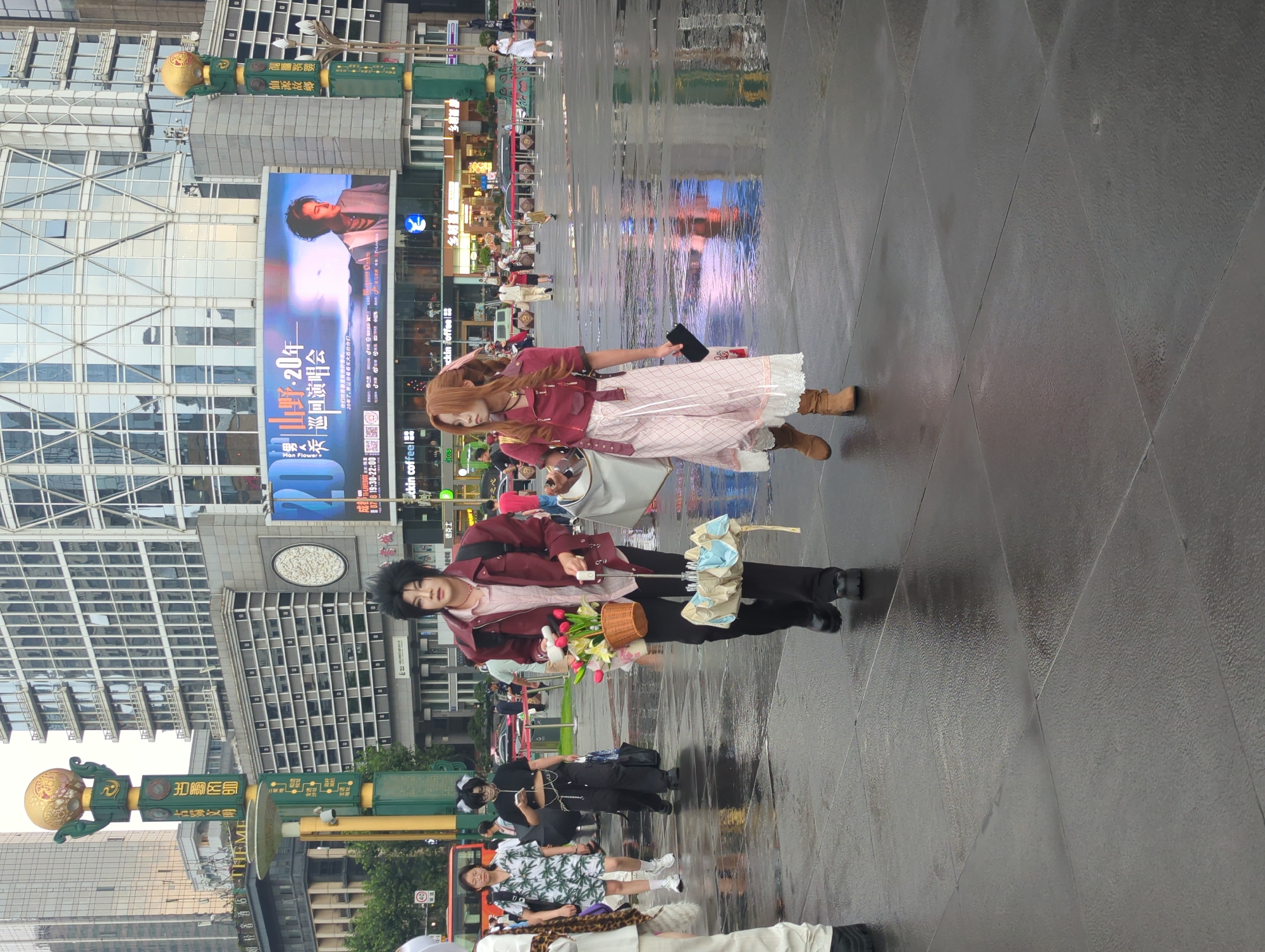 Two individuals, likely cosplayers due to their distinctive attire, are walking across a wet, paved public square. The individual on the left, dressed in a red jacket over a lighter colored shirt and dark pants, carries a basket of flowers and a folded umbrella. The individual on the right wears a reddish-pink top, a patterned skirt, and brown boots. They are walking from left to right in the frame. In the background, large modern buildings with reflective glass facades dominate the skyline. A prominent digital billboard displays an advertisement. Streetlights and other urban infrastructure, including a bus, are also visible. The ground is wet and reflective, indicating recent rain or maintenance. The environment appears to be an urban center, specifically identified as Qingyang District, China.