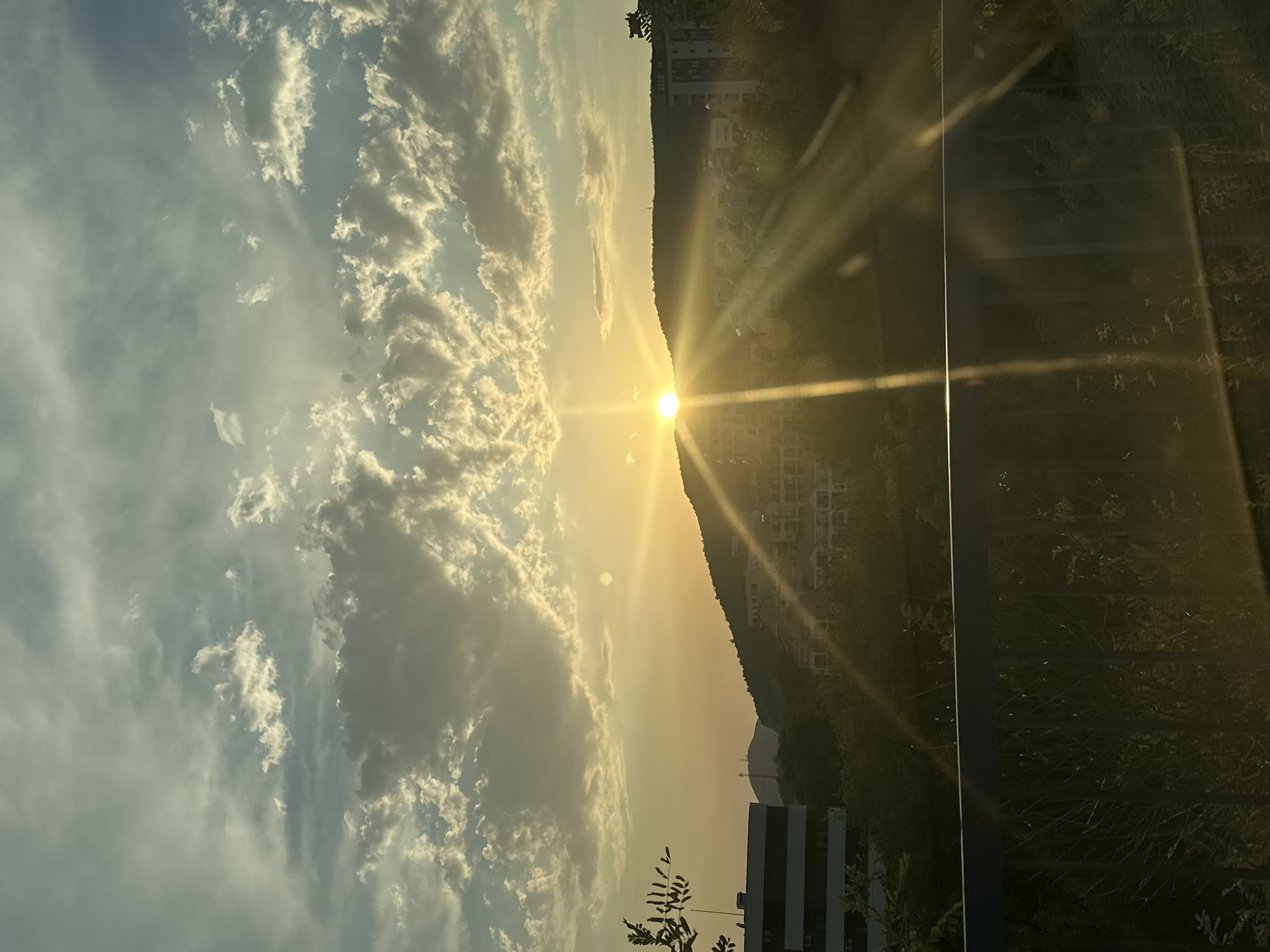 A sunset illuminates the sky over a hillside residential area in Boreti, Montenegro. The sun, a bright orb, is partially obscured by a mountain, casting long, dramatic rays of light that streak across the frame. Clouds, tinged with yellow and white from the setting sun, fill the upper portion of the sky. The hillside is dotted with buildings, some appearing to be apartments or villas, with their facades catching the golden light. In the foreground, a dark railing runs horizontally across the image, with blurred green foliage and shadows visible behind it, suggesting the vantage point is from an elevated position, possibly a balcony or bridge. On the left side of the image, a modern building with horizontal dark bands is visible, its windows reflecting the ambient light. The overall scene conveys a serene, picturesque evening view.