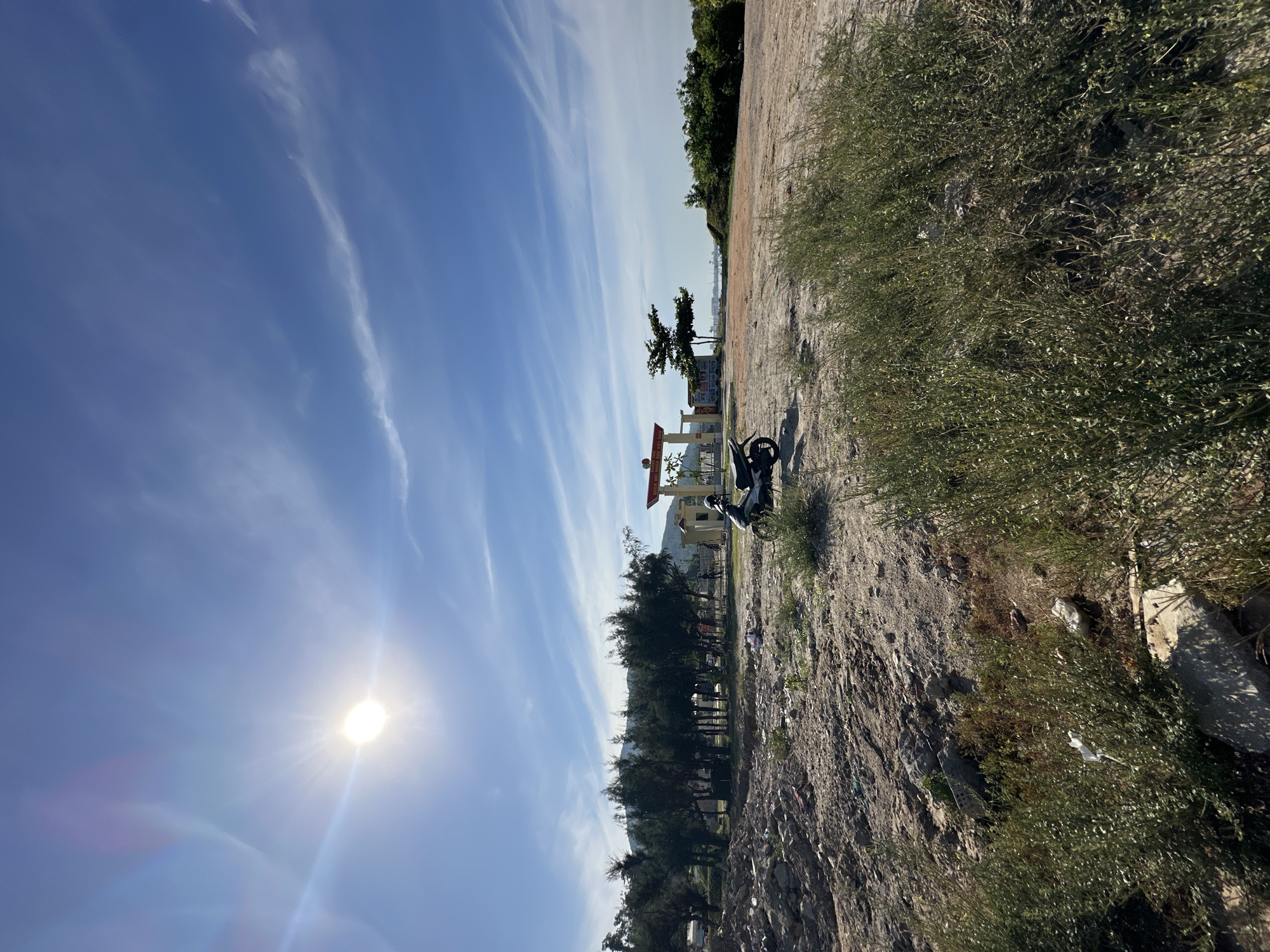 An outdoor, wide-angle, eye-level shot captures a bright, sunny day in Đà Nẵng, Vietnam. The sky is a vibrant blue, dotted with wispy white clouds. The sun is a bright, radiant orb in the upper left quadrant of the image, casting lens flare and illuminating the scene. In the foreground, a patch of rough, uneven ground is covered with sparse, dry brush and scattered rocks. Further back, a line of dark green trees stands tall, with a few buildings and a red-and-white arched structure visible behind them. A black motorcycle is parked on the dirt ground in front of the trees and buildings. The background reveals more greenery and hints of the ocean or a body of water in the distance.