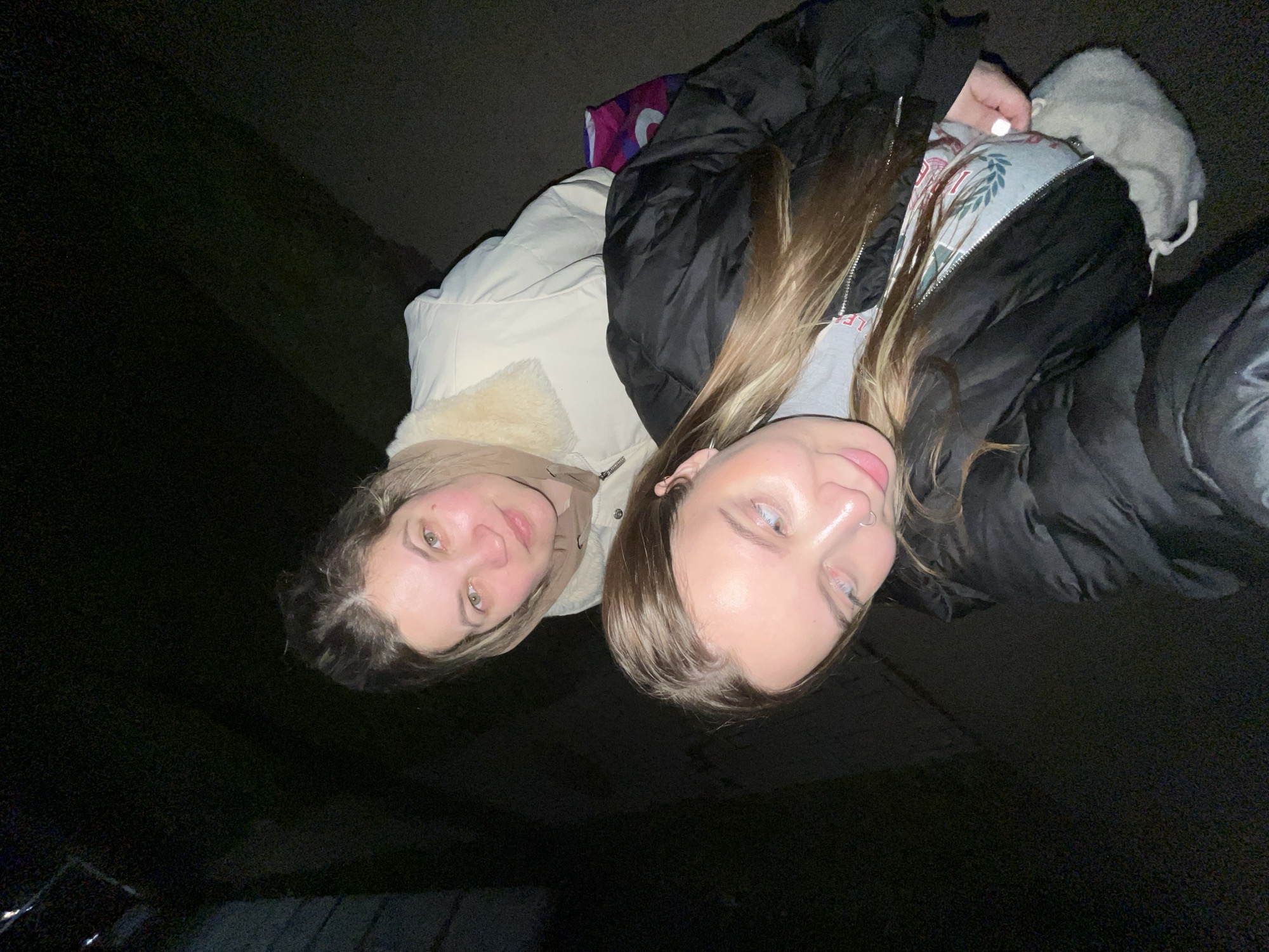 Two young women are standing close together, facing the camera. The lighting is dim, and the background is dark, suggesting it is night or a low-lit environment. The woman on the left is wearing a light-colored jacket with a fur-lined collar. The woman on the right is wearing a black puffer jacket, and her hair is a light brown color. A portion of a gray hooded sweater with a graphic design is visible beneath her jacket. The location is unknown, and there is no information within the photograph that allows for confirming the location of the city of Novopokrovskaya, Russia.