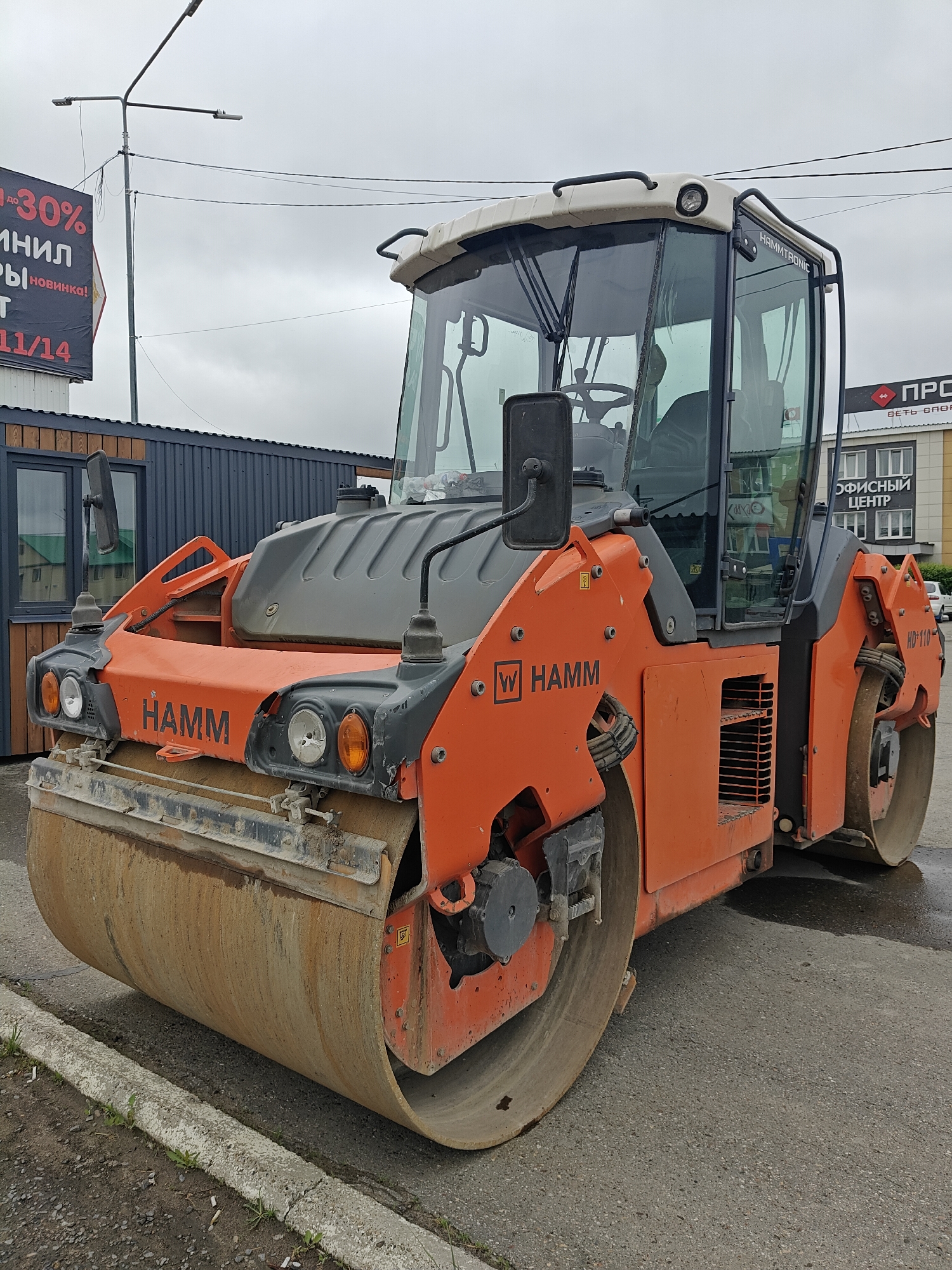 The image features an orange Hamm HD-110 road roller parked on an asphalt surface, near a curb. The roller is oriented with its front drum facing the viewer. Behind the roller are various urban elements, including a small, dark-colored building to the left, a taller building with "ОФИСНЫЙ ЦЕНТР" (Office Center) and the logo of "ПРО СЕТЬ СПOR" on the right. A billboard is seen displaying text in Russian, including "-30%", "ИНИЛ", "Ыы новинка!", and "11/14". A street lamp is visible above the buildings. The sky appears overcast. This photograph was taken in the city of Chaikovsky, Russia.