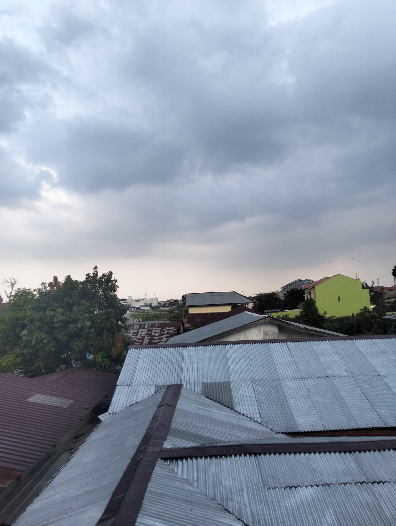 The image depicts a view of rooftops and the sky in the city of Medan, Indonesia. The foreground is dominated by corrugated metal roofs, some appearing weathered with rust. A large, leafy green tree is visible on the left, obscuring part of the view of the buildings behind it. Further in the distance, several structures are visible, including a bright lime green building and other houses with various colored roofs. A few taller buildings, possibly apartment complexes or commercial structures, can be seen in the background. The sky is overcast with heavy, gray clouds, suggesting an impending storm or a dull, cloudy day. The overall scene conveys a densely populated urban environment with a mix of residential and possibly some institutional buildings.