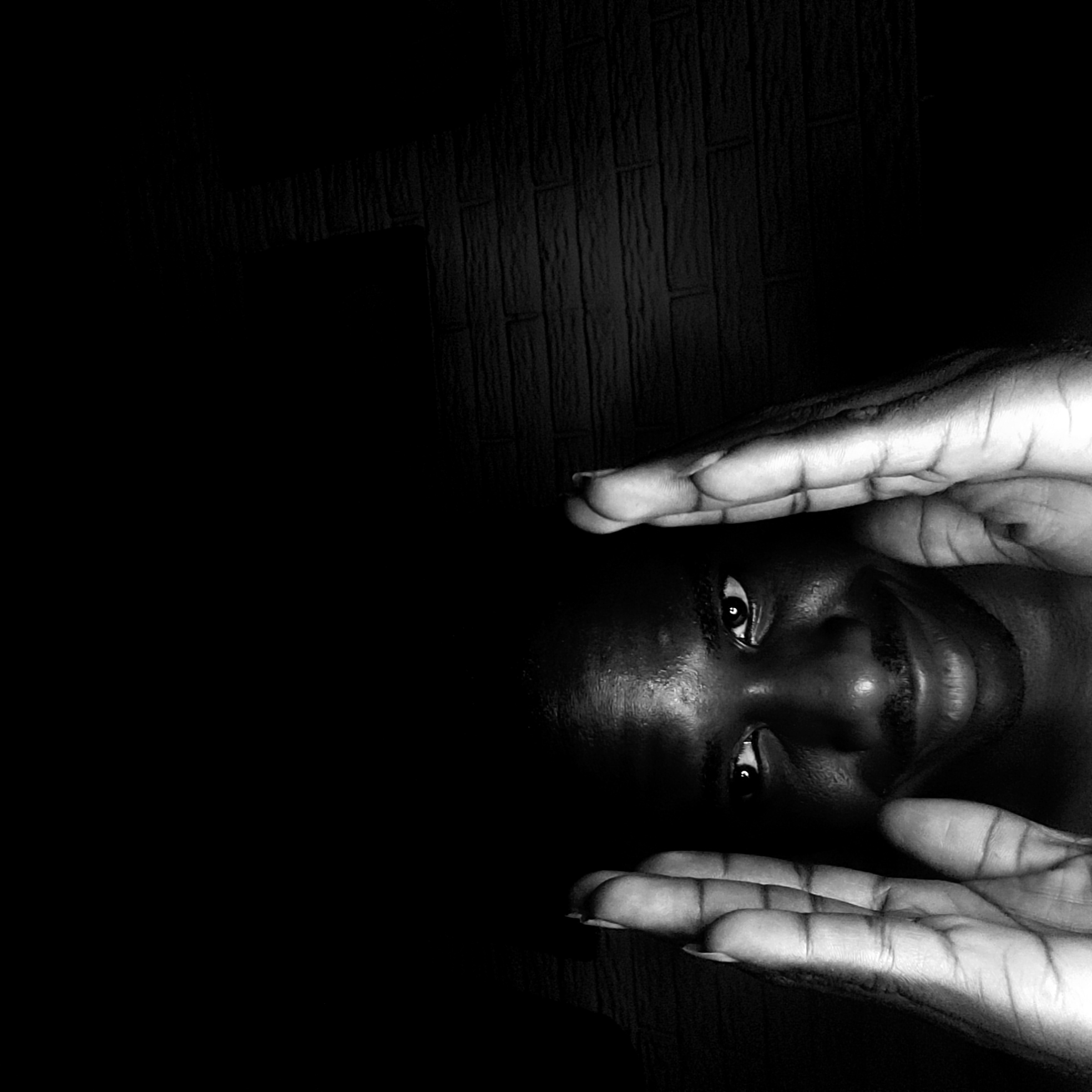 A black and white, close-up, eye-level shot shows a person with dark skin lying down and looking up. The person's face is framed by their own hands, which are positioned above and below the face. The background appears to be a dark, textured wall, possibly made of wood or a similar material. The setting is unidentified, and there is no information to confirm the location as Narayi, Nigeria.