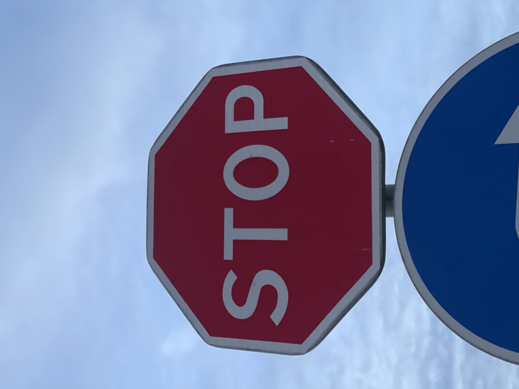A red octagonal stop sign with white lettering spelling "STOP" is mounted on a pole above a blue circular sign with a white arrow indicating a right turn. The sky behind the signs is a pale blue with scattered clouds. The scene is outdoors, likely on a street or roadway, given the presence of traffic signs. The angle of the photograph is from below, looking up at the signs and the sky. The lower portion of the blue sign is visible, revealing part of a white arrow.