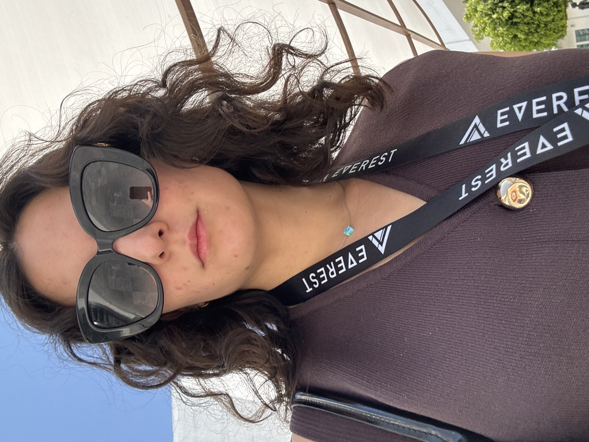A medium-close-up photograph taken outdoors features a woman from the upper chest level, centered in the frame. The subject has dark, curly hair and wears large, dark, cat-eye style sunglasses, which reflect a smartphone in the right lens. Her skin is light, displaying minor facial blemishes. Her lips are closed. She is attired in a dark brown, possibly ribbed, sleeveless garment. Around her neck, she wears a thin silver chain with a small, light blue, square-shaped pendant. A black lanyard with the white text "EVEREST" and a white triangular logo is draped around her neck. A circular, gold-toned pin is affixed to her garment on the left side, beneath the lanyard. A dark strap, consistent with a purse or bag, is visible over her left shoulder. The background features a clear blue sky on the upper left and a large, white canopy structure with visible framework on the right, indicating an outdoor, shaded area. A blurred green bush is visible in the lower right background. The overall lighting suggests a bright, sunny environment. The precise location within the city of Puebla, Mexico, cannot be identified from the visual details provided in the image.