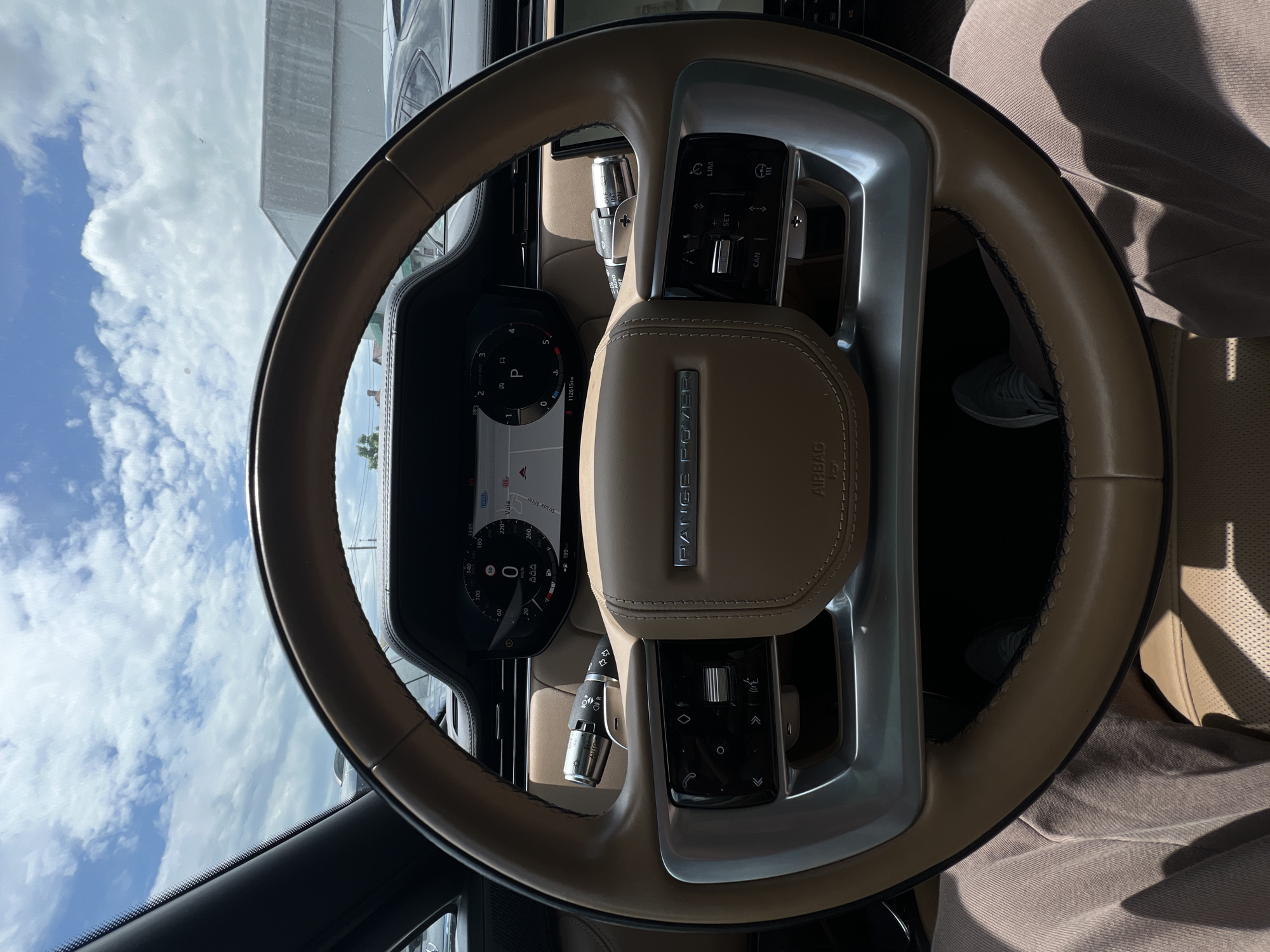A first-person view from the driver's seat of a Range Rover, looking at the steering wheel and dashboard. The steering wheel is tan leather with silver accents and controls for the infotainment system and cruise control. The digital dashboard displays speed, navigation, and other vehicle information. Outside the car, a bright sky with scattered clouds is visible, along with buildings and other vehicles. The interior of the car appears to be luxurious, with leather seats and modern technology. The image was taken in Cluj-Napoca, Romania, on a sunny day.
