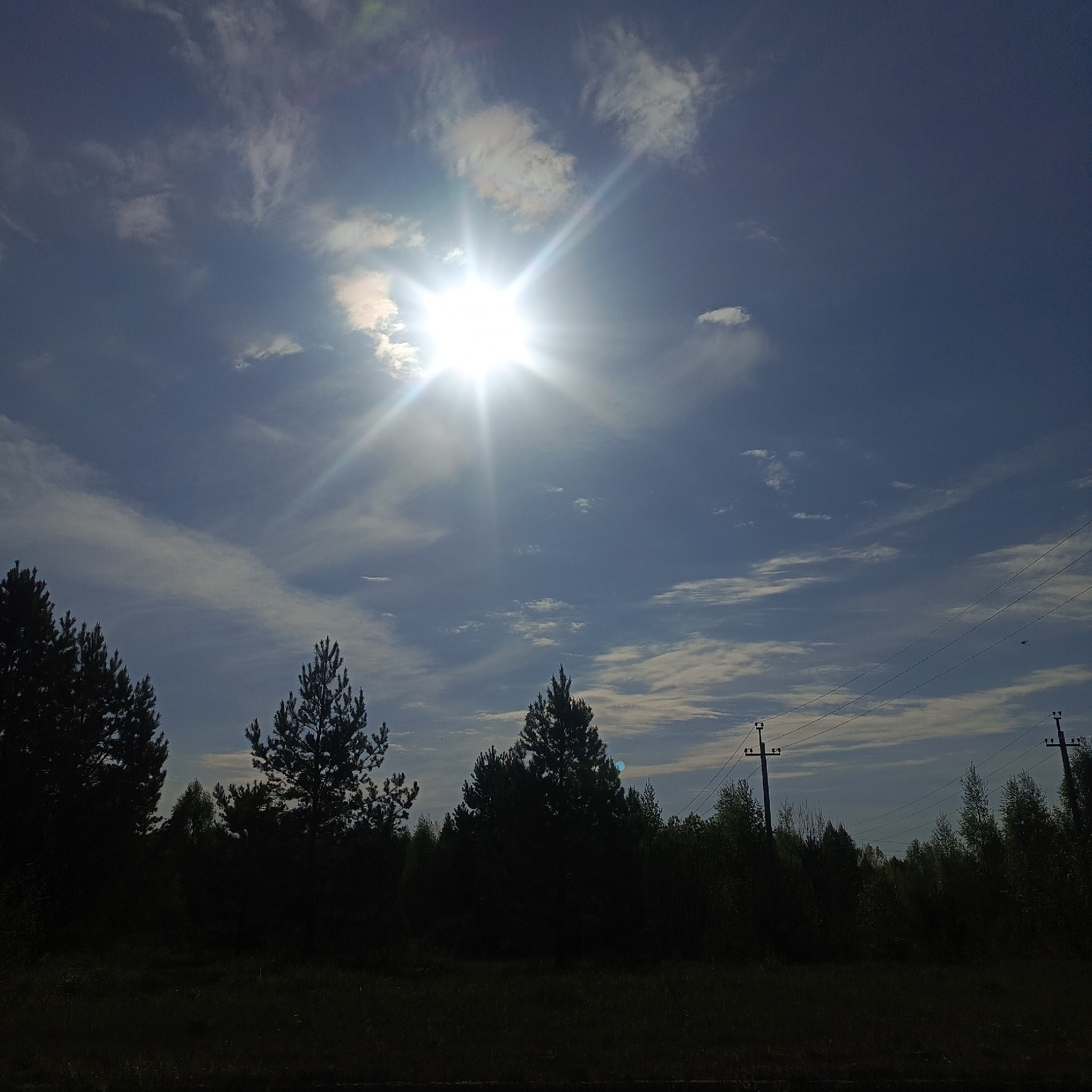The image shows a daytime scene in Марковский, Russia. The sun is high in the sky, partially obscured by thin clouds, with visible sunbeams. The sky is blue with wispy white clouds scattered throughout. In the foreground, silhouetted trees and vegetation line the horizon. Power lines and utility poles are visible to the right, extending into the distance. The foreground is mostly dark, suggesting a low vantage point or shadow.