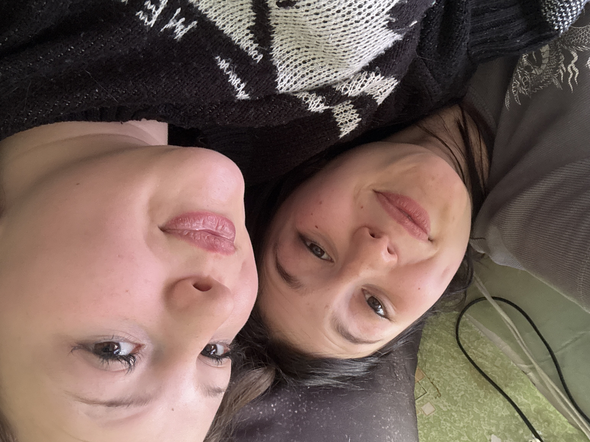 Two women are lying down close together, looking at the camera. The woman on the left has fair skin and brown eyes. She is wearing a black and white patterned sweater. The woman on the right has darker hair and is wearing a gray shirt. The background is a light green surface with wires visible. The location in the city of Фоки, Russia, is not discernible from the image.