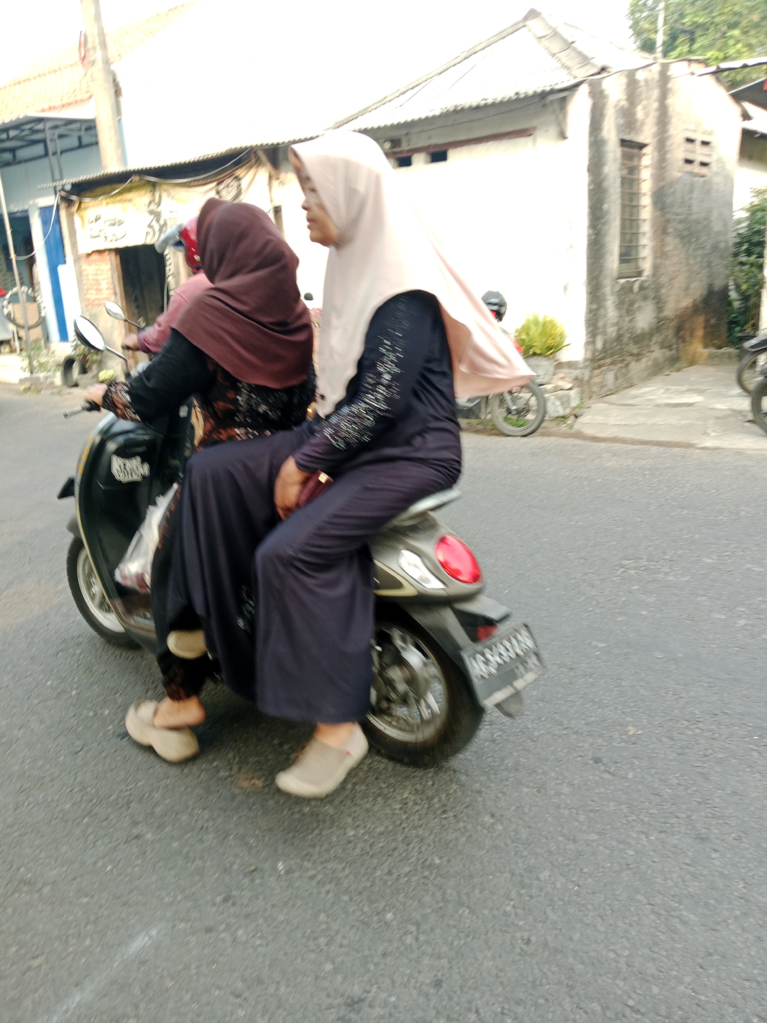 Two women are riding a dark colored scooter on an asphalt road. The woman in the front is driving, wearing a maroon hijab and a patterned shirt underneath a dark colored outer garment. The woman in the back is wearing a light colored hijab and a dark colored dress. Both women are wearing casual shoes. The scooter has a red taillight and a license plate with illegible white characters on a black background. In the background, there is a building with a corrugated metal roof and a white wall, as well as some vegetation and another scooter parked to the left. The scene appears to be in a residential area within the city of Kediri, Indonesia.