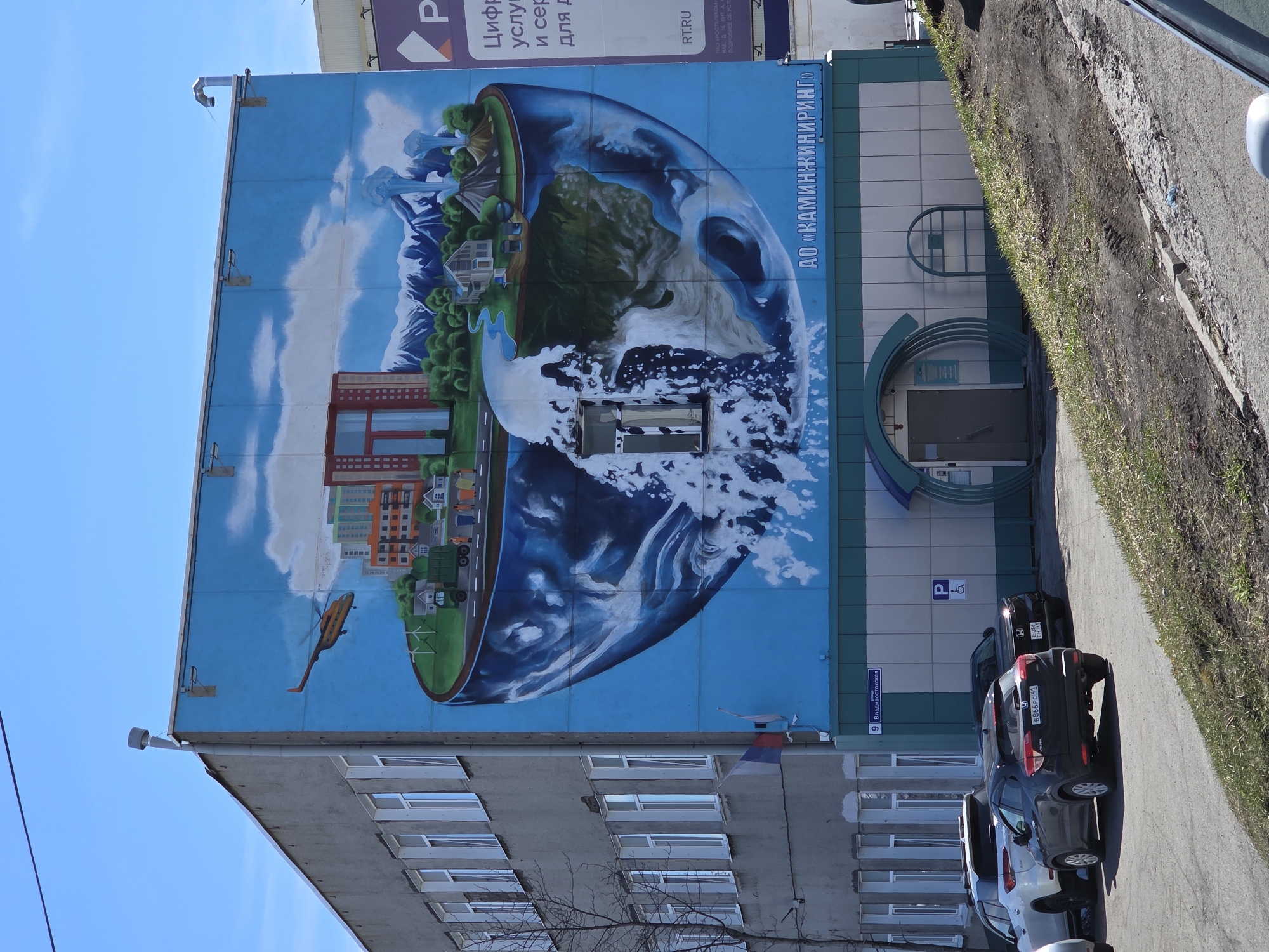 A vertically oriented photograph captures a building with a striking mural of a cross-section of the Earth, combined with cityscapes and nature scenes. The mural occupies a significant portion of the building’s facade, depicting mountains, water features, and urban structures within the Earth's curved shape. A helicopter is painted in the lower-left corner of the mural.
The building has multiple rows of windows beneath the mural. Adjacent to the mural is a section of the building with a parking sign and a distinct teal and white tiled design, incorporating a circular doorway.
Several cars are parked along the side of the building, with a street sign reading "Владивостокская" visible, indicating the location. The OCR text shows the name "АО «КАМИНЖИНИРИНГ»" suggesting this may be the name of the company located in the building. The presence of the street name "Владивостокская" suggests a location in or near Vladivostok, Russia. Additional text mentions "РОСТЕЛЕКОМ," a Russian telecommunications company, further supporting this.