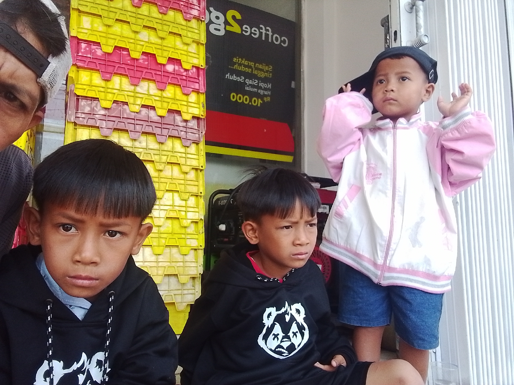 In the city of Cianjur, Indonesia, two young boys are photographed in front of a stack of yellow and pink plastic crates. The boy on the left, wearing a black hoodie with a white graphic of a stylized panda, is looking directly at the camera with a serious expression. He has short, dark hair and appears to be about 8-10 years old. To his right, another boy, younger than the first, is wearing a pink and white jacket and blue shorts. He has a dark baseball cap on backwards and is looking to his left with his hands raised slightly. Behind the boys is a window with text that appears to be in Indonesian, including the word "Coffee." A person's head, wearing a black baseball cap, is visible in the upper left corner of the frame, partially obscuring the scene. The lighting suggests it is daytime.