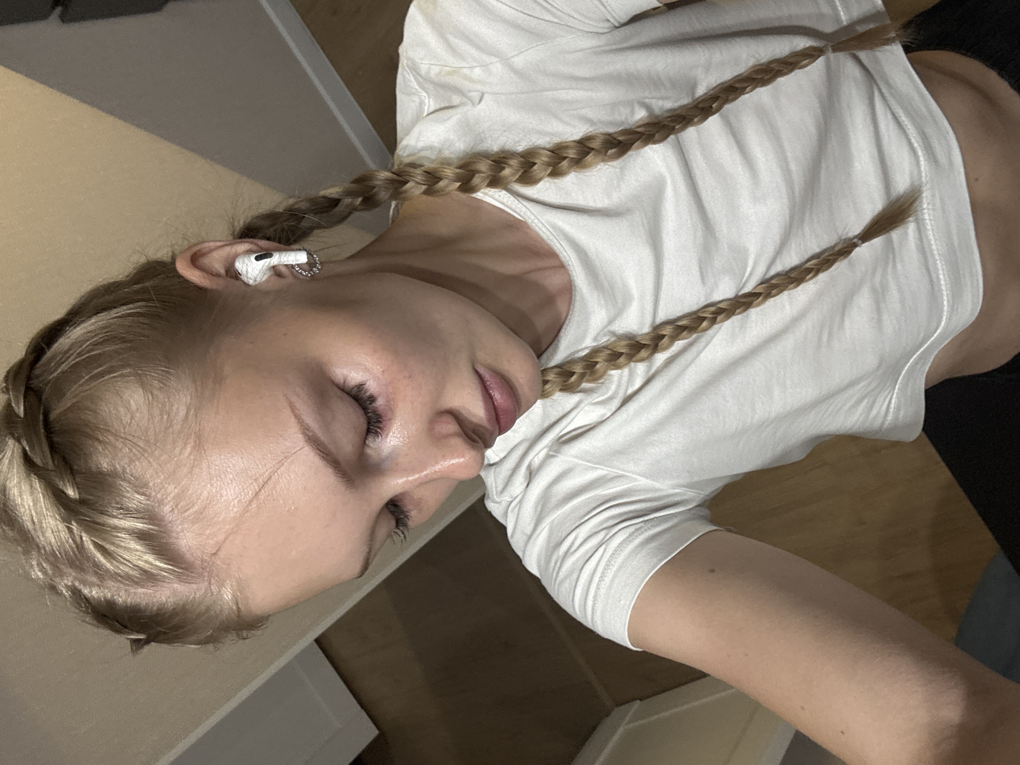 A young blonde woman with light skin and freckles is shown from the chest up, looking down and to her left. She has two braids that fall over her shoulders, one on each side. She is wearing a white t-shirt and has white earbuds in her ears. The background appears to be an indoor setting with wooden floors and a doorway visible. There is no indication of the city of Perm, Russia, in the image.