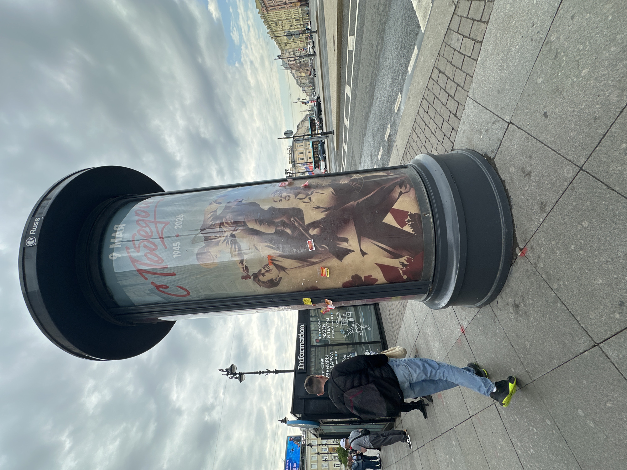 Here's a detailed description of the image:
**Setting:** An outdoor scene, likely a street in Saint Petersburg, Russia. The sky is overcast with gray clouds.
**Objects:**
* A large cylindrical advertising column or kiosk with a poster commemorating the Victory Day (May 9th, 1945-2026), a Russian holiday celebrating the end of World War II. The poster features figures resembling soldiers and related imagery. The column has a black top and base. There are small stickers on the column.
* An "Information" sign.
* Street lighting poles and other urban furniture visible in the background.
* Buildings lining the street in the distance.
* Cars are visible in the distance.
**People:**
* At least three individuals are present in the image.
* One man wearing a black jacket, blue jeans, and distinctive black and neon green sneakers. He is walking by the information sign. He carries a black backpack.
* Other people are walking nearby in the background.
**Actions/Interactions:**
* People are walking on the sidewalk.
* The man in the black jacket and blue jeans is walking.