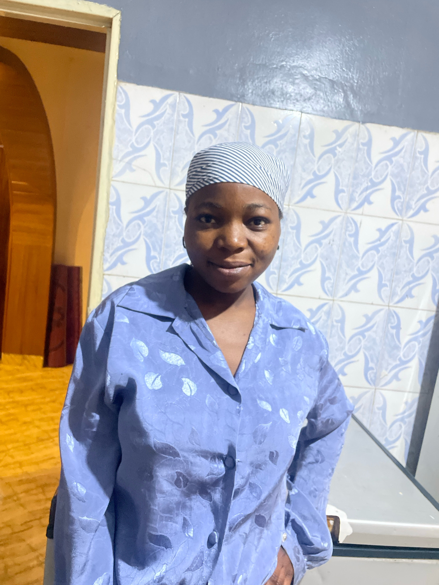 A medium shot shows a Black woman wearing a blue, patterned long-sleeved shirt and a striped headscarf, standing in what appears to be a kitchen in Jos, Nigeria. The woman is looking directly at the camera with a slight smile. Behind her, a tiled wall features a blue and white pattern. To her left, a doorway with an arched opening reveals a yellow, textured floor. The scene is illuminated by artificial light, creating shadows and highlighting the textures of the environment and the woman's clothing.