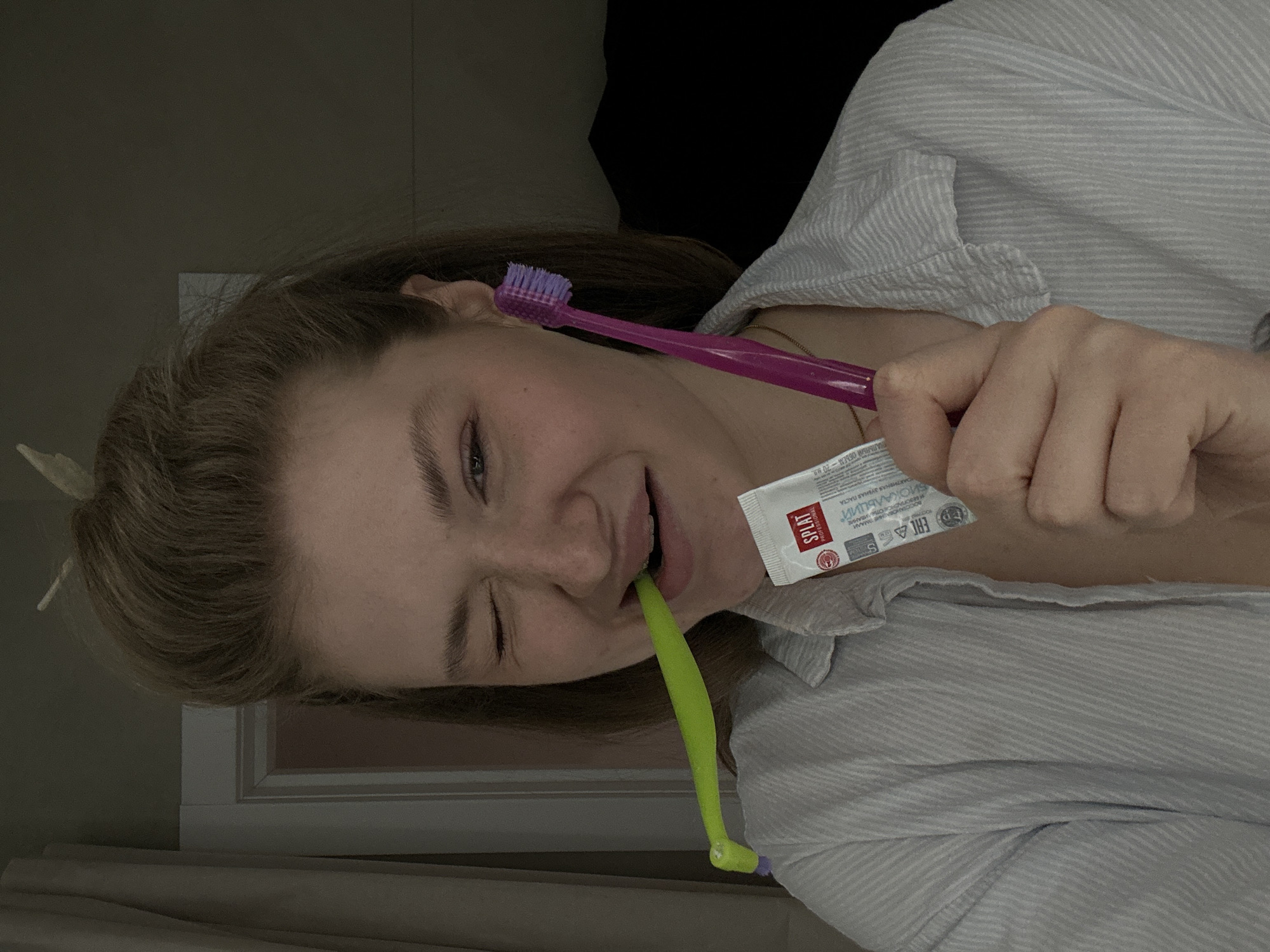 The image shows a woman indoors. She is holding a purple toothbrush and a tube of "SPLAT" brand toothpaste in her hand. She has a green dental brush in her mouth. The tube of toothpaste indicates it is "BIOKALCIY" and that it is "SAFE WHITENING". The text on the tube also indicates "ACTIVE TOOTHPASTE". The toothpaste is also marked with "PROFESSIONAL". It also says on the tube, that it is for "ENAMEL" and "SAFE WHITENING". The woman is wearing a striped collared shirt and is winking one eye. The location is not specified, but text on the toothpaste tube is written in Russian. The full address for Kazan, Russia is not specified in the image.