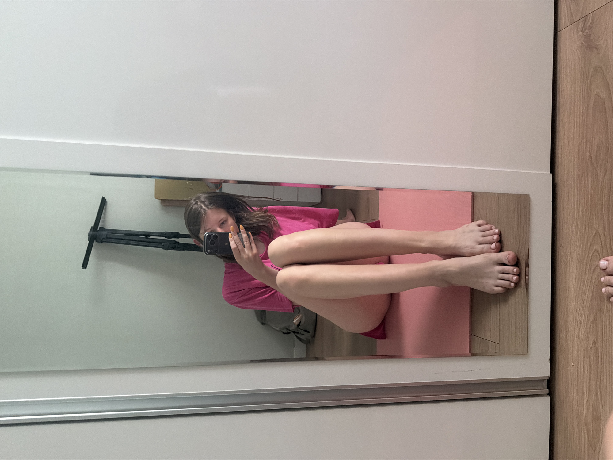 A young woman is taking a selfie in a full-length mirror, sitting on a pink yoga mat in what appears to be a bedroom or studio. She is wearing a bright pink t-shirt and has her legs crossed, with her bare feet visible. The mirror reflects a tripod in the background, a backpack, and a portion of the room's interior. The floor is made of light-colored wood. The scene is lit with natural light, suggesting it is daytime. The woman's face is partially obscured by her phone as she takes the photo. Her fingernails are painted an orange-yellow color.