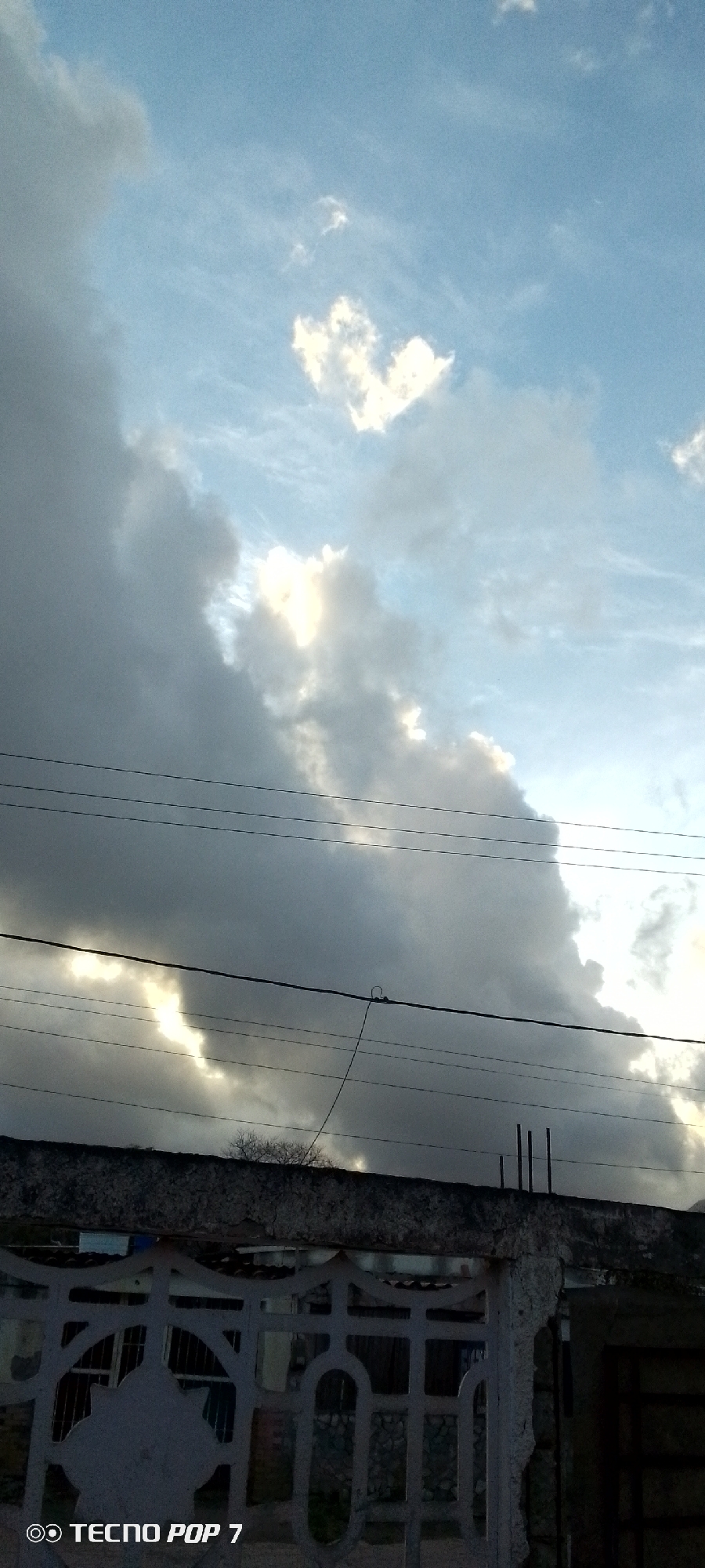 The image shows a cloudy sky in Altagracia, Venezuela. The sky is mostly blue with some white and gray clouds. The sun is shining through the clouds, creating a bright spot in the center of the image. In the foreground, there is a fence, a low wall, and some power lines.